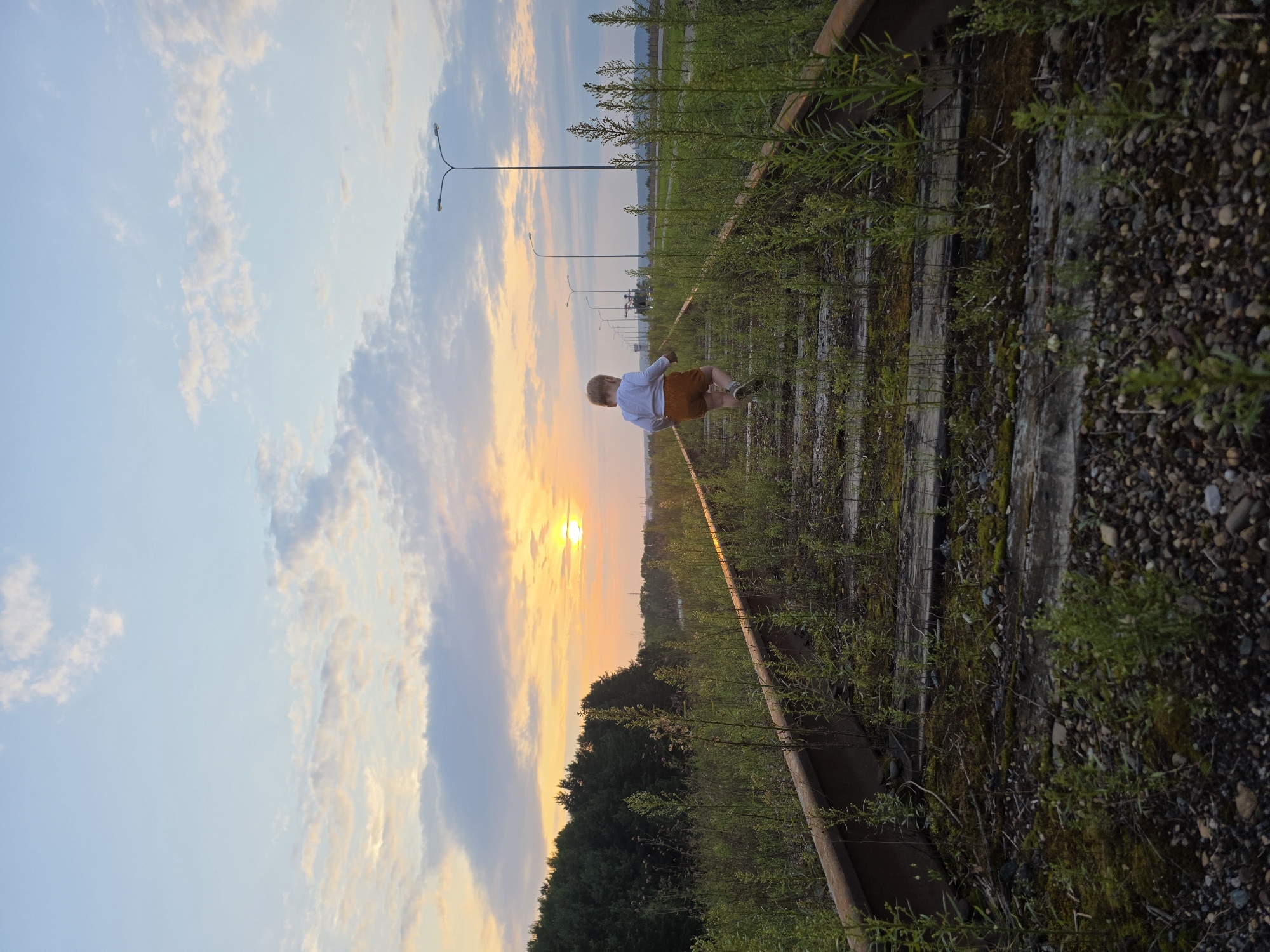 A child with blonde hair, wearing a white long-sleeved shirt and orange shorts, walks away from the camera on abandoned train tracks. The tracks are overgrown with weeds and grass, with wooden sleepers visible beneath. In the background, a line of streetlights stretches into the distance, and beyond that, a forest can be seen. The sun is setting, casting a warm glow over the sky, with clouds tinged with orange and pink. The scene appears to be in an industrial or undeveloped area of Chaikovsky, Russia, possibly near the Kama River embankment, given the presence of old railway lines that were once used for industrial purposes. The lighting suggests it is late afternoon or early evening.