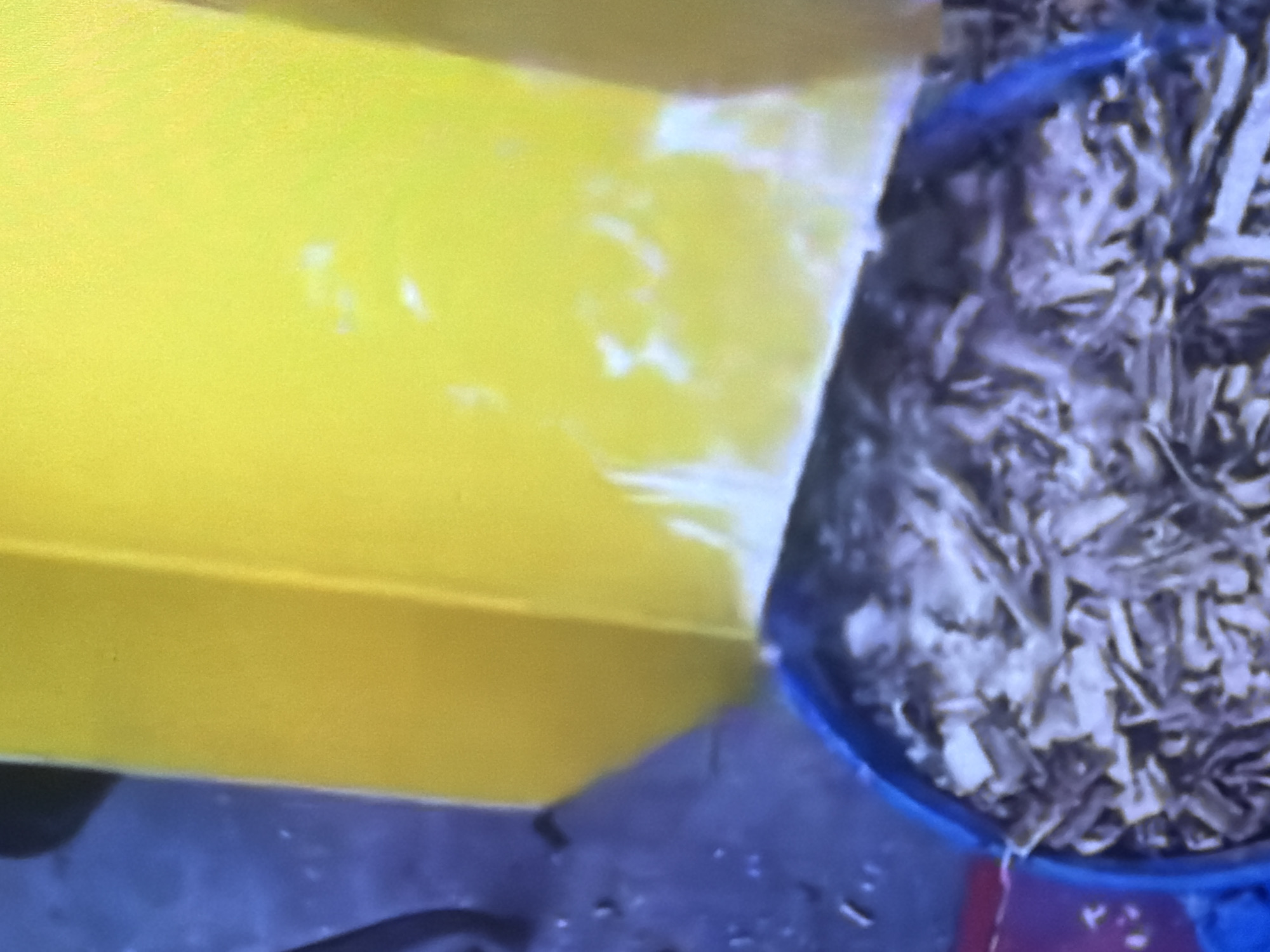 A close-up shot of a yellow object above a blue bin filled with shredded material. The blue bin appears to be a bucket or a large container. The shredded material inside the bin is a mix of light brown and grey pieces, resembling wood shavings or shredded paper. The yellow object is bright and appears to be made of plastic or painted metal. The background is blurred and shows a dark blue surface, possibly a floor or ground. There are some white smudges on the lower part of the yellow object, and a few small dark specks on the blue surface. The lighting is diffused, suggesting an indoor or shaded outdoor setting. There are no people or animals visible in the image. The location is identified as Bukuru, Nigeria.