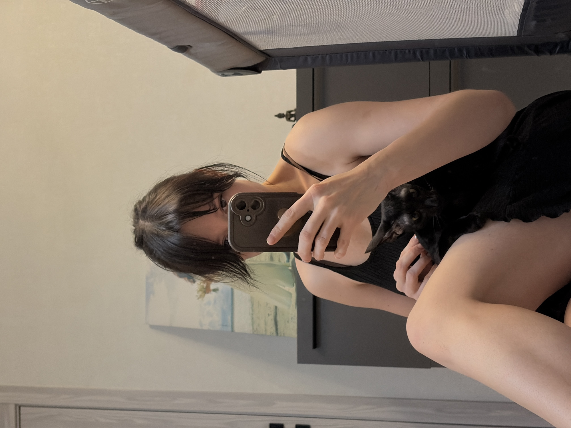 A woman with dark hair and bangs is taking a selfie in a room. She is holding a phone in her right hand, obscuring part of her face. A small, black cat is nestled against her chest. She is wearing a black top. The background includes a gray piece of furniture, a painting on the wall, and a white cabinet. The location in the city of Chaikovsky, Russia, cannot be determined from the image.