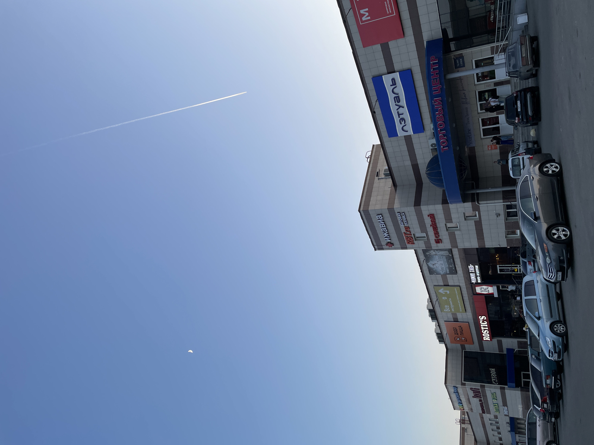 Here is a detailed description of the image:
The image shows a street scene in Izhevsk, Russia. The sky is clear and blue with a contrail from an airplane, and a crescent moon is visible. On the right side of the image is a multi-story shopping center. Signs on the building are in Russian and include: "Торговый центр л'этуаль," which translates to "Shopping Center L'Etoile" (a beauty store); "Рижавиа," (Rizhavia) a local appliance store; "Rostic's," a restaurant chain; "Все инструменты," meaning "All Tools"; "Додо Пицца," a pizza chain; and "Мамин Хлеб," meaning "Mom's Bread". Several cars are parked along the street next to the shopping center. People can be seen walking in the entrance to the shopping center.