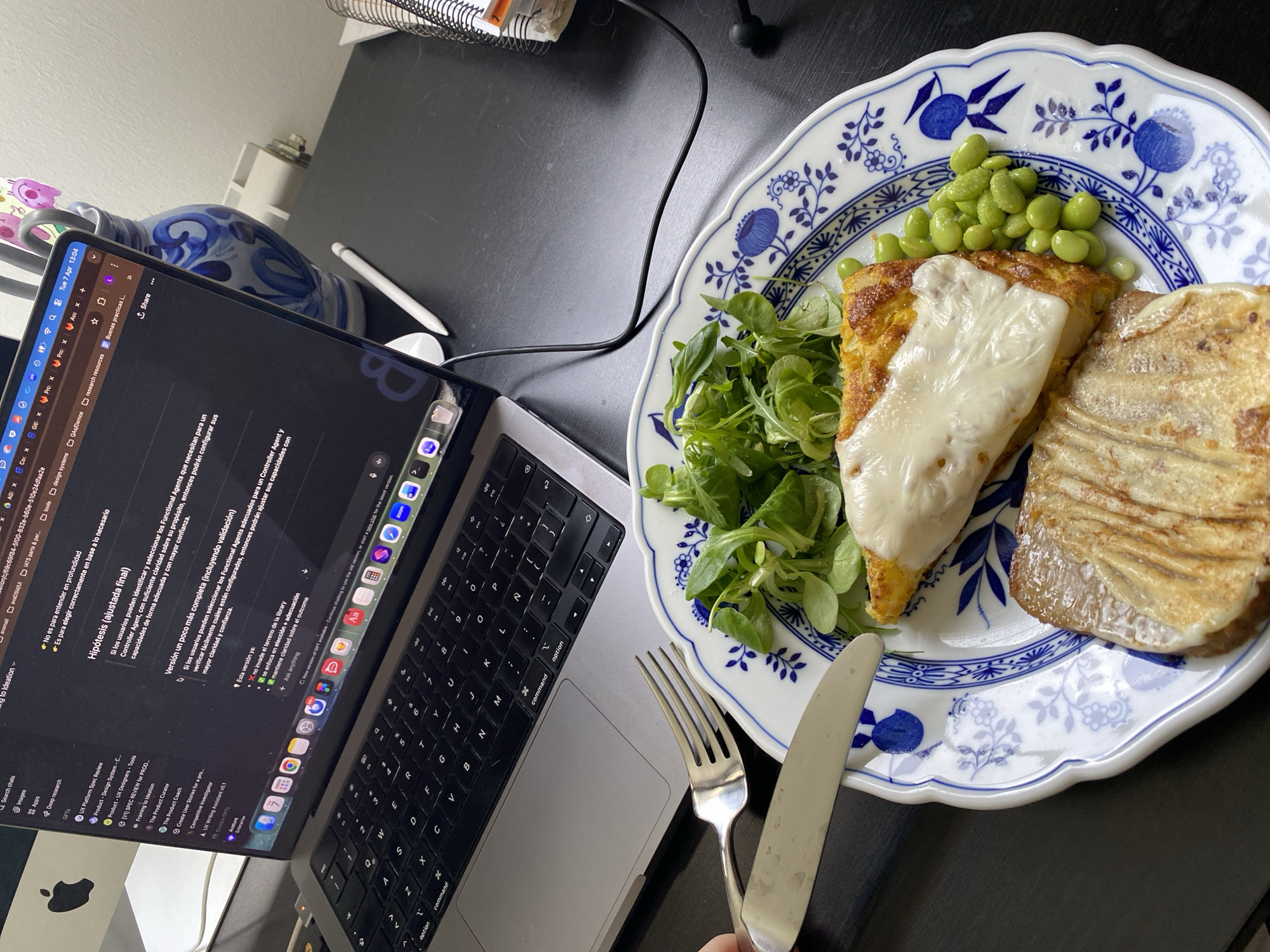 This image shows a laptop and a plate of food on a dark surface, likely a desk or table. The laptop screen displays text in Spanish, suggesting someone is working or studying. The plate contains a meal consisting of some salad greens, what appears to be edamame, a cheesy slice and a breaded and grilled piece of something. A fork and knife are positioned near the plate, seemingly ready for use. The scene suggests a person is having a working lunch or meal break while using their laptop. The image does not provide any information about the city of Villaviciosa, Spain.