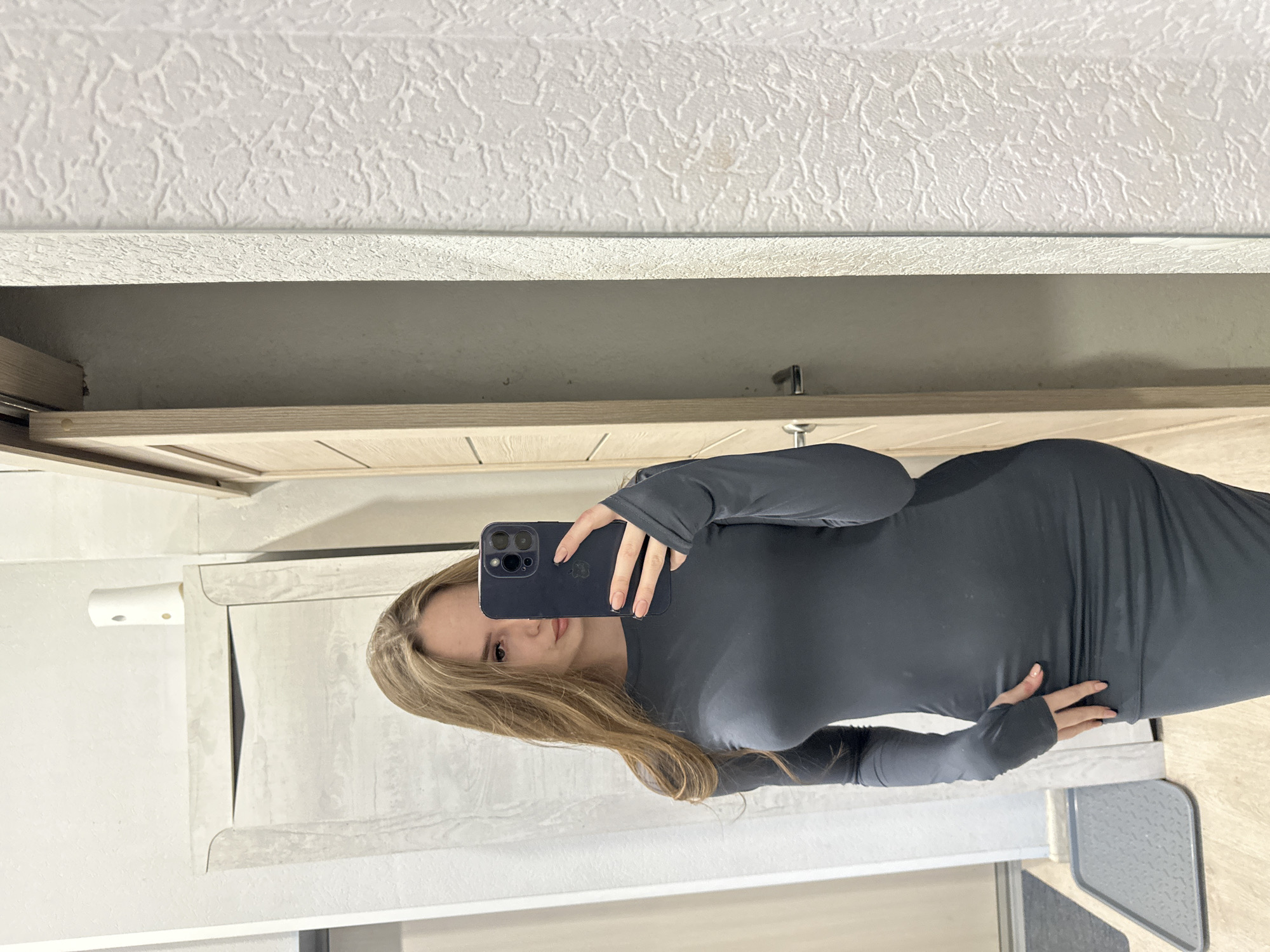 A woman is taking a selfie in a bathroom. She is wearing a form-fitting gray long-sleeved dress. She is holding a black iPhone in front of her face. Her long, light-brown hair flows over her shoulders. The bathroom has light-colored wood cabinets and a textured white ceiling. There is a toilet paper holder on the wall. The location is Unknown, Unknown.