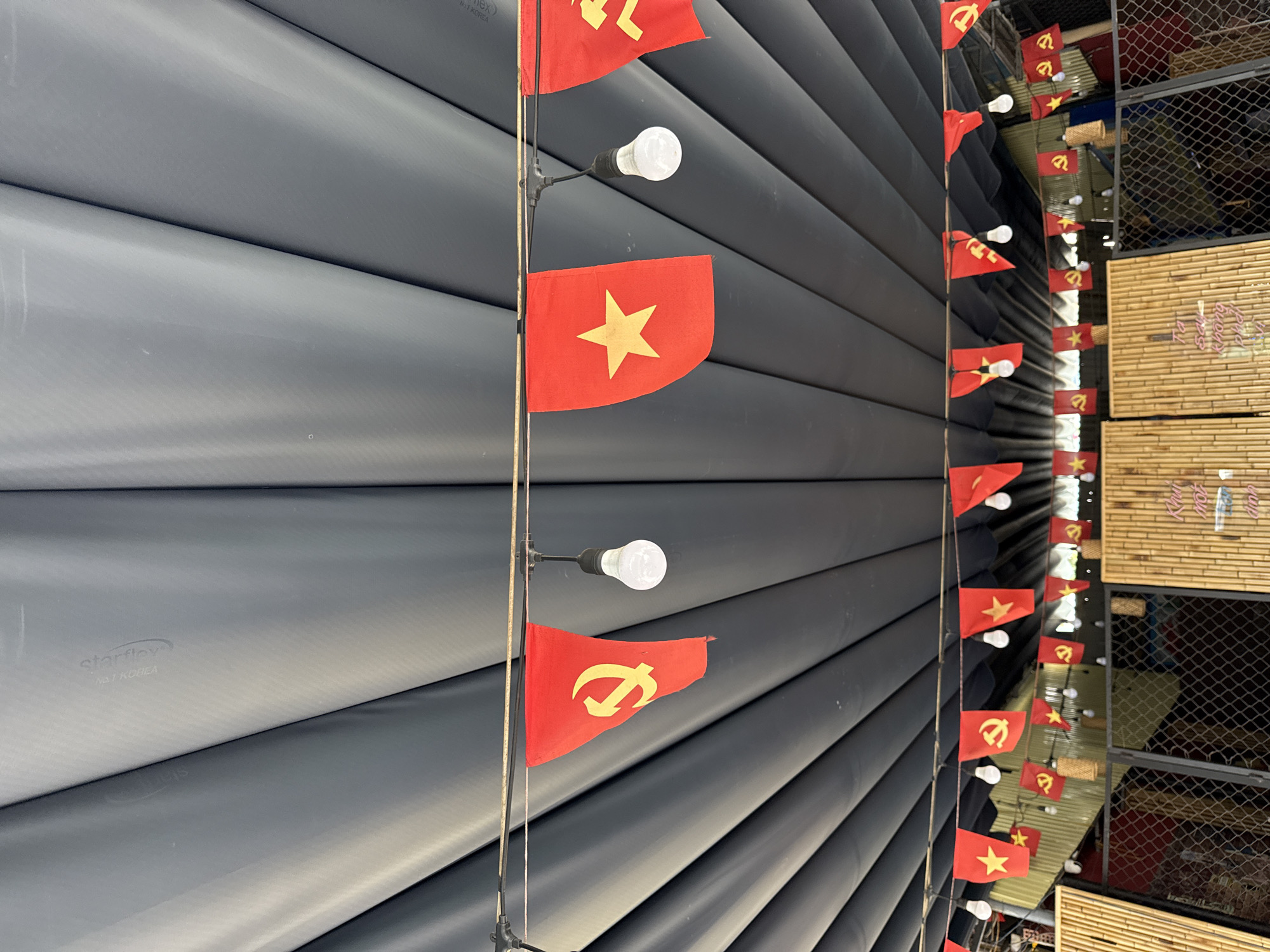 The image presents a view of a structure decorated with a series of small red flags, seemingly alternating between the flag of Vietnam (with a yellow star) and another flag with a hammer and sickle. These flags are strung along a vertical line, interspersed with small light bulbs. The backdrop features a wall composed of long, parallel, gray, tube-like structures, possibly ventilation ducts or pipes. To the right, there is a lattice-like structure with bamboo-like elements, possibly a fence or wall. The scene is captured in a tight, medium shot with an angle, and is possibly located in the city of Đà Nẵng, Vietnam, based on the flags.