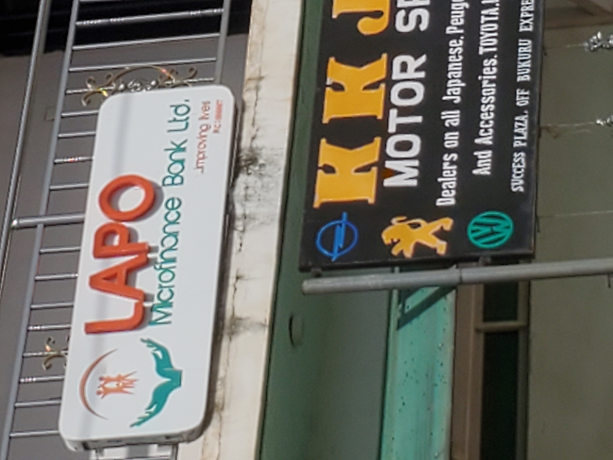 The image shows two signs. The upper sign is white with red and green lettering. The red lettering says "LAPO" and the green lettering says "Microfinance Bank Ltd.". Below this, in smaller print, it says "Improving lives". The lower sign is black with yellow and white lettering. The yellow lettering says "KK J MOTOR SP". Below this, in white lettering, it says "Dealers on all Japanese, Peugeot And Accessories. TOYOTA.H.". At the bottom of the black sign, it says "SUCCESS PLAZA. OFF BUKURU EXPRE". The signs are mounted on a building. The building has a metal railing in front of it. The environment appears to be an outdoor urban setting.