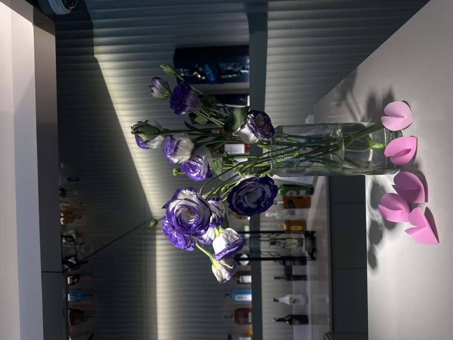 A vase with blue and white flowers sits on a white surface next to pink paper cutouts. The background features a modern interior with horizontal blinds and a shelf stocked with various bottles, possibly liquor. The lighting is bright, casting shadows and highlighting the textures of the flowers and paper cutouts. The scene is simple, yet the color contrast and lighting create a visually appealing composition.