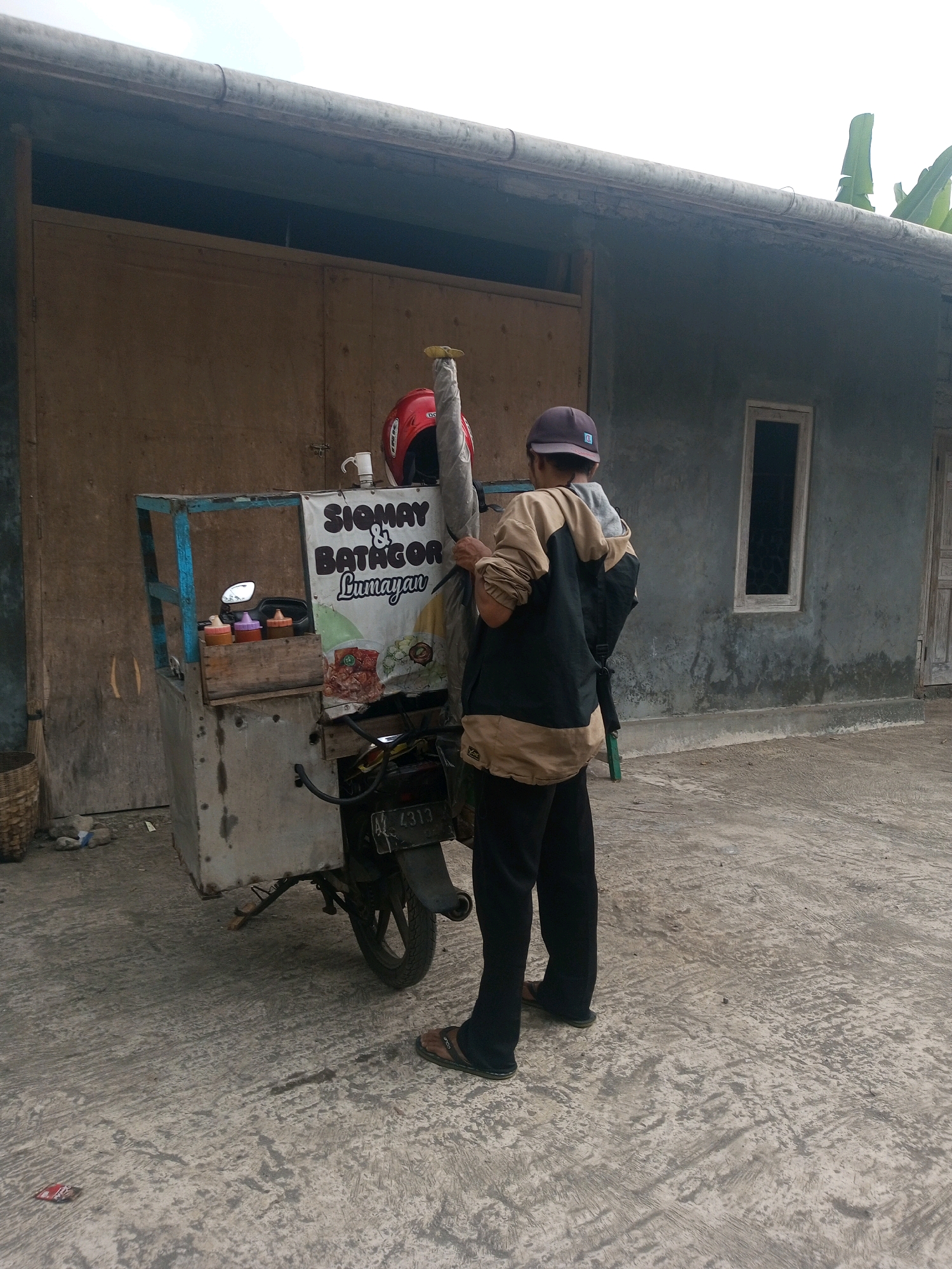 A person stands near a food cart attached to a motorcycle in Krasak, Indonesia. The food cart has a blue frame and a grey metal body, with a white banner advertising "SIOMAY & BATAGOR Lumayan." On the cart, there are three bottles of sauce and a motorcycle mirror. A red helmet rests on top of the cart. The person is wearing a tan and black jacket, black pants, and dark sandals, with a purple cap on their head. The motorcycle license plate is visible and reads "4313". The scene takes place on a concrete surface in front of a building with a large wooden door and a small rectangular window. The overall environment appears to be an outdoor, urban setting.