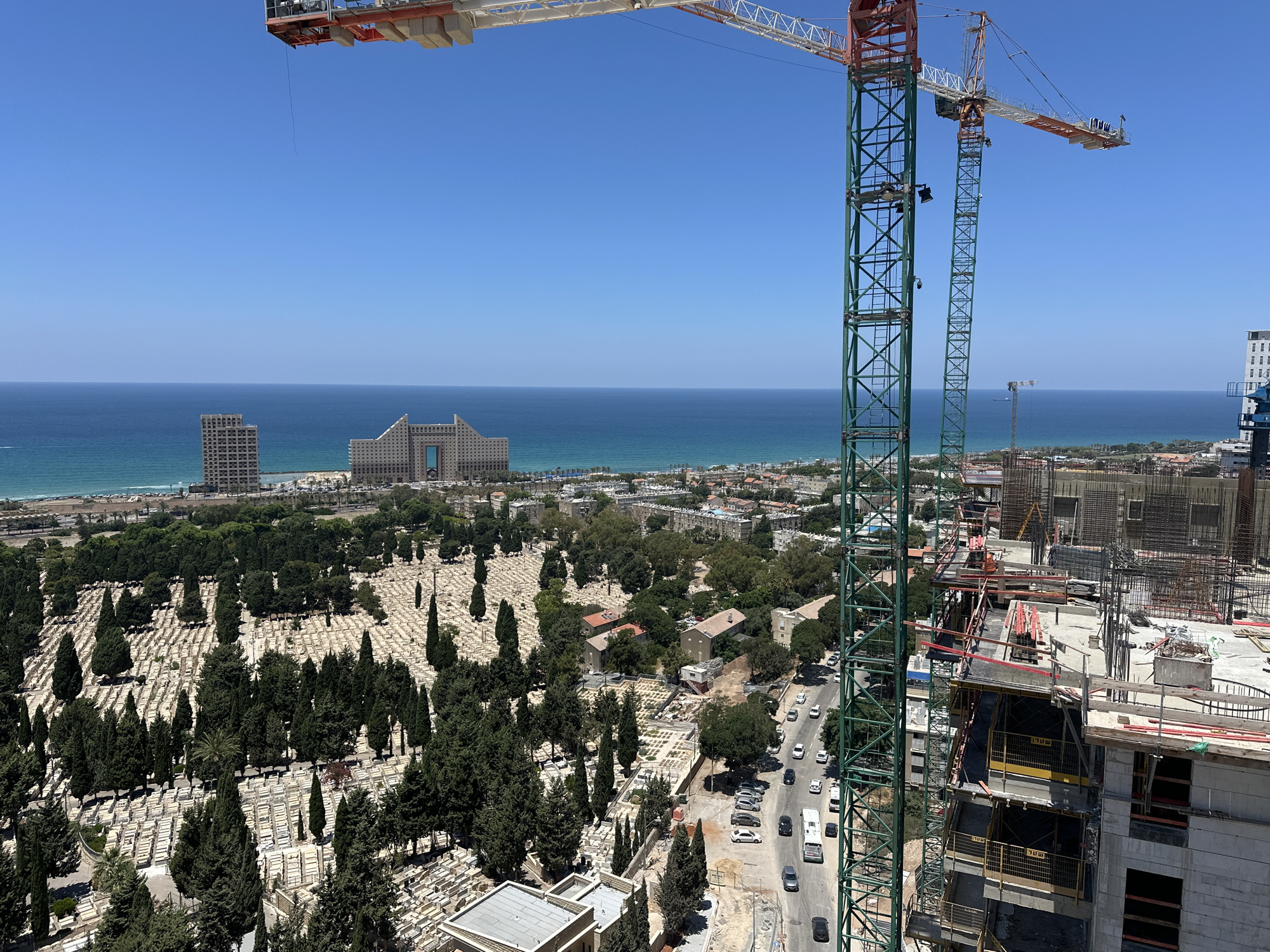 An aerial view captures a construction site in Haifa, Israel, with two large green and orange cranes dominating the foreground. The cranes are positioned next to a partially built high-rise structure, its concrete frame and rebar visible. Below the construction, a dense, sprawling cemetery with rows of white headstones and numerous evergreen trees stretches across the landscape. Beyond the cemetery, a segment of the city is visible, featuring residential buildings, roads with a few cars, and lush greenery. In the distance, the deep blue Mediterranean Sea meets the clear, bright blue sky. Two distinct buildings, one a tall, modern high-rise and another with a unique, angled architectural design, stand prominently near the coastline.