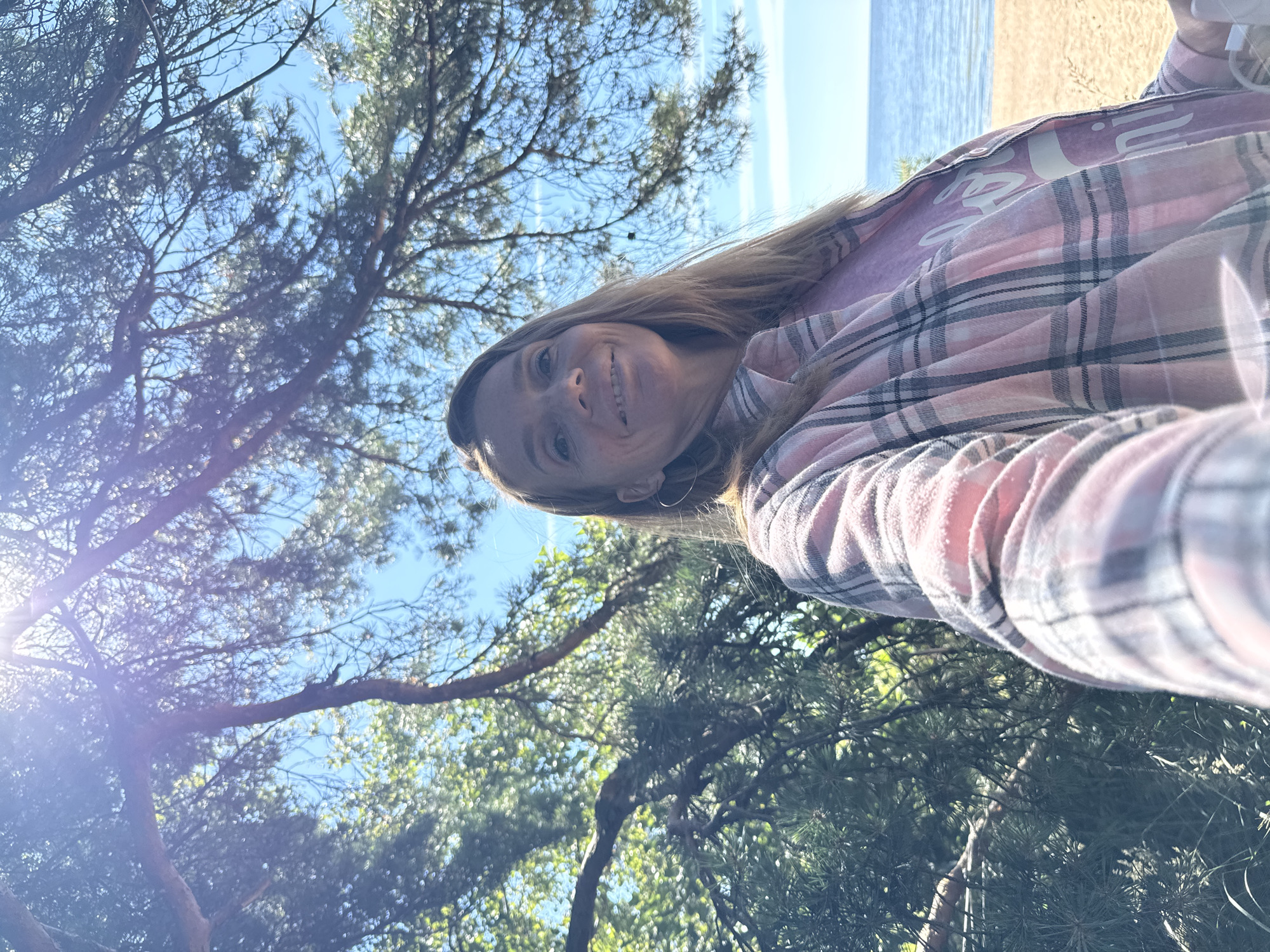 A woman with blonde hair and a wide smile takes a selfie outdoors, with a body of water and a sandy beach visible in the background. She is wearing a pink and gray plaid shirt over a purple t-shirt with white lettering. Sunlight streams through the branches of a pine tree, casting a lens flare across the top of the image. The location is identified as Zelenaya Roshcha, Russia.