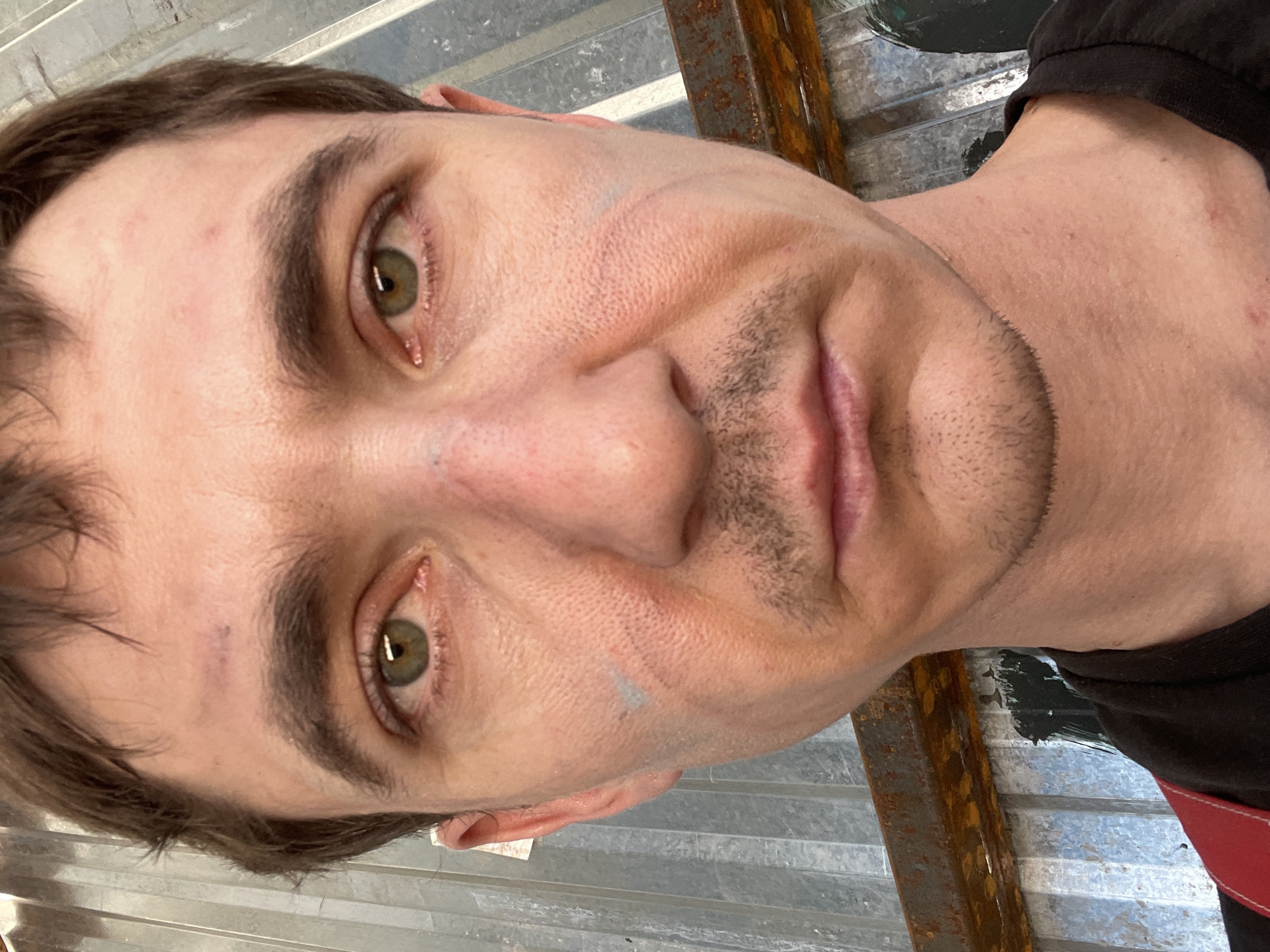 A close-up, selfie-style photograph captures a Caucasian male, appearing to be in his late 20s or early 30s, against a corrugated metal background. The man has light brown hair, parted and somewhat disheveled, with a receding hairline. His eyes are a striking green, and his skin is fair with visible pores and some redness, particularly around the nose and cheeks. He has a light mustache and stubble. There are faint smudges of blue or grey paint on his cheeks, one on the left and one on the right, and a bruise or mark on his forehead above his left eyebrow. He is wearing a black t-shirt with red suspenders visible. The background consists of vertical, grey corrugated metal sheets, with a rusty metal beam running horizontally across the lower portion of the frame. The lighting is natural, casting soft shadows. The scene is outdoors, possibly in an industrial or construction area. The photographer appears to be standing outdoors on a sunny day. The location is the city of Sokury, Russia.