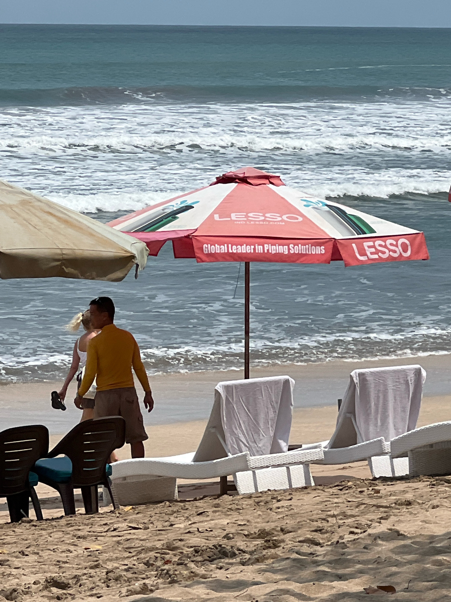 The image depicts a beach scene in Kuta, Indonesia, with the ocean and sky in the background. In the foreground, there are beach chairs and umbrellas. A man in a yellow long-sleeved shirt and brown shorts is walking with a woman in a white top, holding a sandal in his hand. An umbrella with the branding "LESSO Global Leader in Piping Solutions" is prominent in the center of the frame. The beach is sandy, and the ocean has moderate waves.