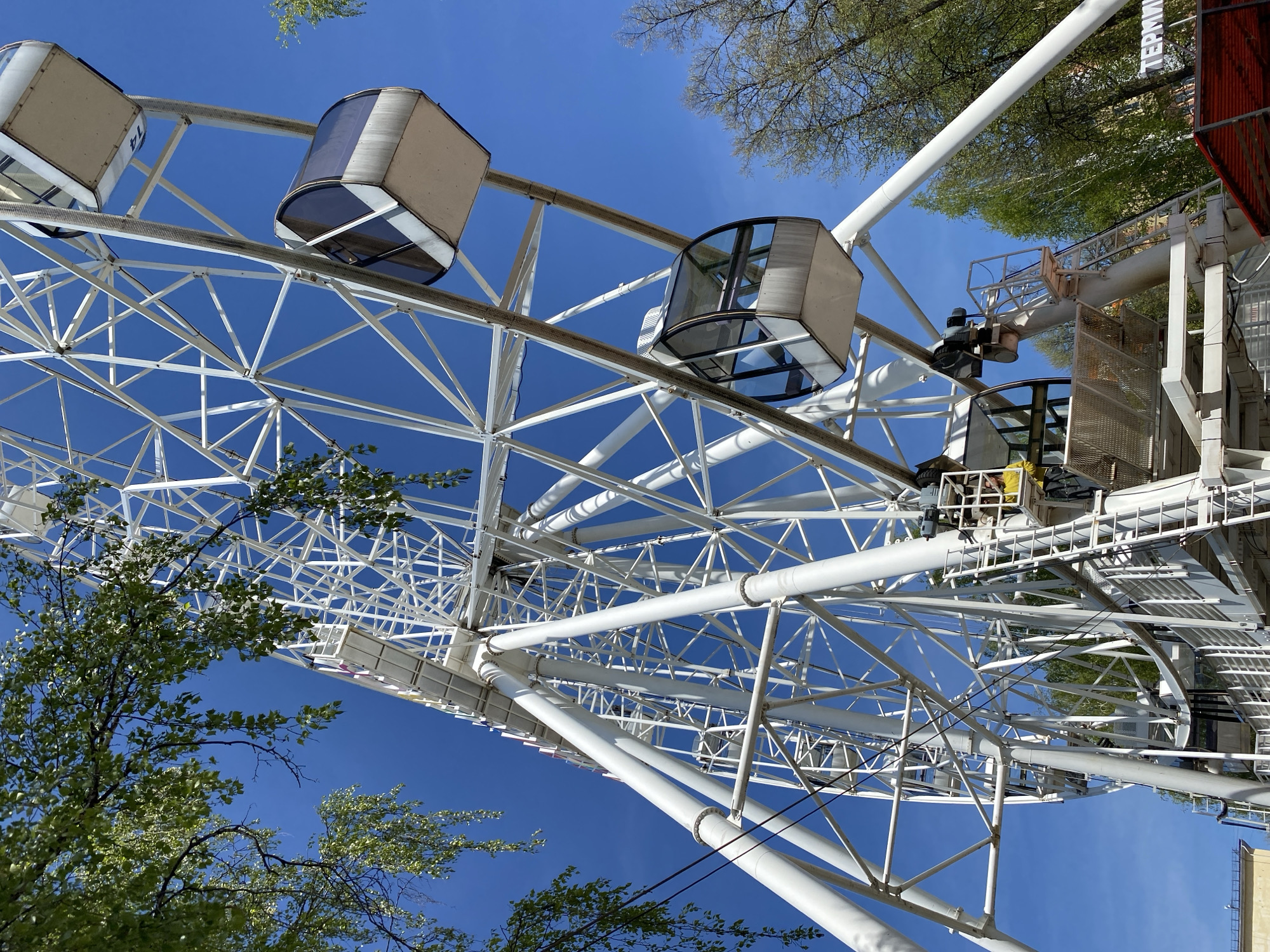 An eye-level, low-angle shot depicts a white Ferris wheel against a bright blue sky. The wheel's gondolas are enclosed with partially tinted windows. The Ferris wheel appears to be located in a park, as there are trees with green foliage visible around the structure. The city is Izhevsk, Russia.