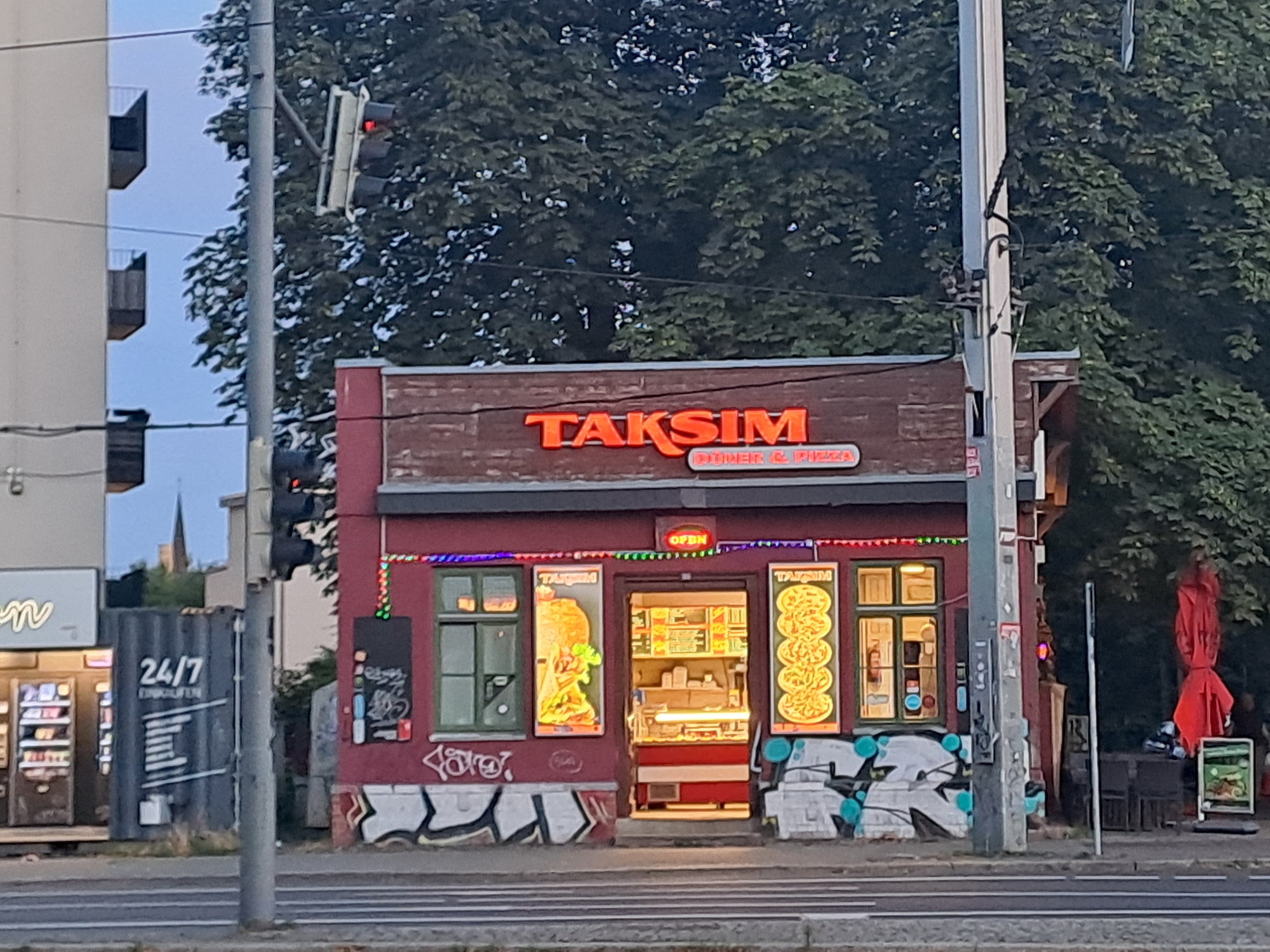 The image depicts a street-level view of "Taksim" restaurant, a kebab and pizza establishment, located in Leipzig, Germany. The building is a narrow, two-story structure painted maroon with a brick-like facade on the upper portion. Neon lights, including a red "OPEN" sign, illuminate the storefront. The main signage features the name "TAKSIM" in orange neon letters above the words "DÖNER & PIZZA".
The ground floor has a central entrance with a brightly lit interior displaying food items and a counter. To the left of the entrance, a green-paned window displays a large image of a döner kebab. To the right, another green-paned window showcases an illuminated advertisement for pizza. The lower part of the building is adorned with graffiti.
In front of the restaurant, a traffic light is visible on a pole, with the red light illuminated. Adjacent to the restaurant on the left is a dark, corrugated metal container with "24/7" printed on it, along with additional text and lines of information. Further to the left, a portion of a white building with balconies is visible.
To the right of the restaurant, an outdoor seating area with chairs and tables is partially obscured by a large red umbrella. A smaller green sign with food imagery stands near the seating area. The scene is set against a backdrop of dense green trees, indicating an outdoor urban environment. The street in the foreground has multiple lanes marked by white lines. The overall impression is that of a typical street scene in a European city, likely during daylight or early evening.