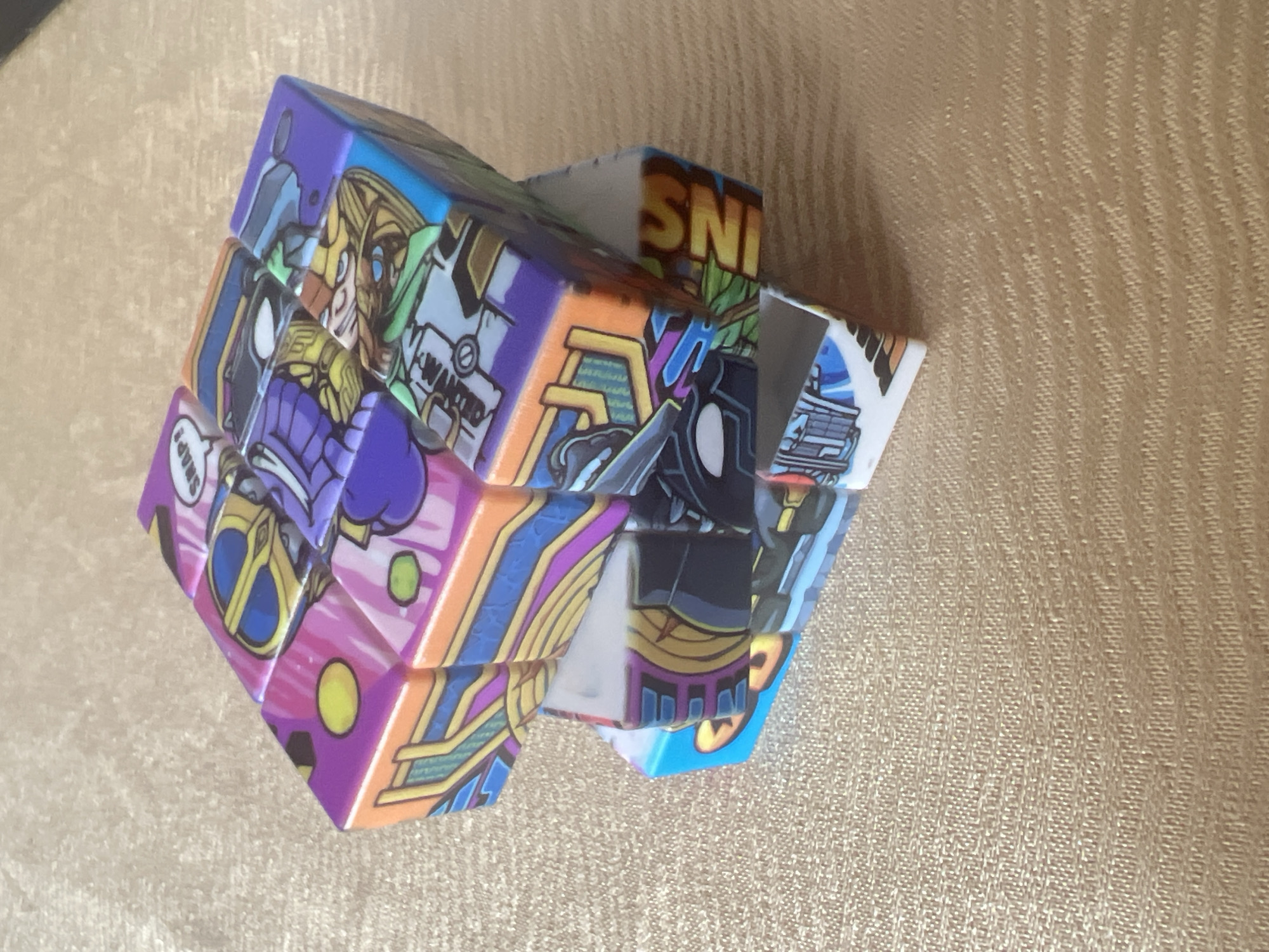 A partially disassembled, custom-designed Rubik's Cube-like puzzle rests on a light brown, textured surface. The puzzle consists of multiple cubic segments, some interconnected in an irregular formation, and one piece partially detached from the main structure. Each face of the segments features colorful, stylized illustrations. Visible imagery includes a yellow-gold helmet with blue goggles, a purple-armored character with a "SNAP!" speech bubble, a blonde-haired character wearing green, a character with a black mask and white eyes, and a blue and white character with intricate geometric designs. The partial text "SNIP" is visible on one piece, alongside solid yellow and green circular motifs on purple and pink backgrounds, respectively. The underlying surface displays a fine, consistent woven or textured pattern. The specific geographic location in the city of Burgas, Bulgaria, cannot be determined from the visual content of the image.