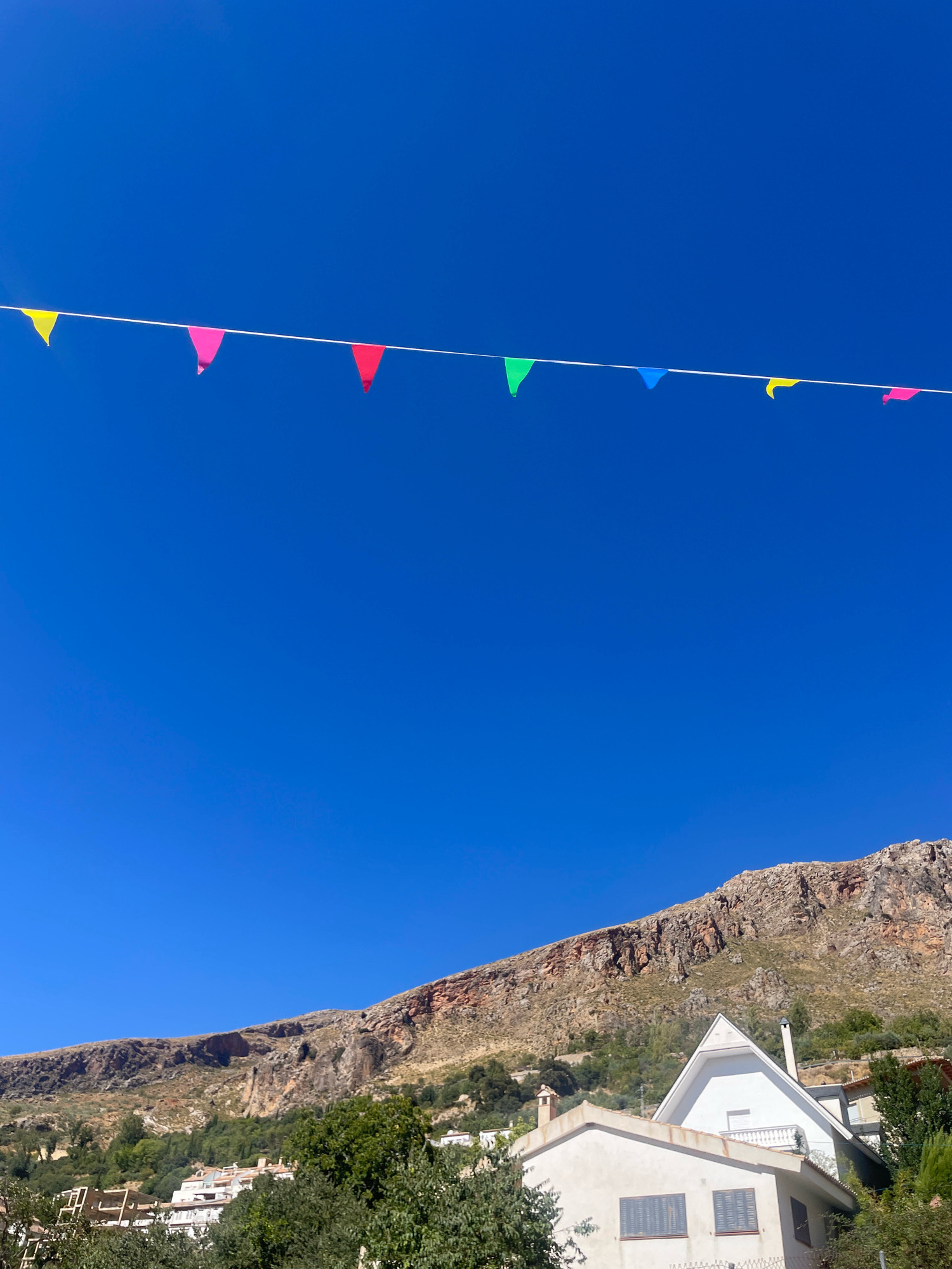 The image shows a clear blue sky above a mountainous landscape in Güéjar Sierra, Spain. A string of colorful triangular flags, including yellow, pink, red, green, and blue, are strung across the sky. Below the flags, a rocky mountain slopes downwards, covered in sparse vegetation. Several white buildings with terracotta roofs are visible nestled amongst trees on the lower part of the mountain and in the foreground. The scene appears to be set during the daytime.