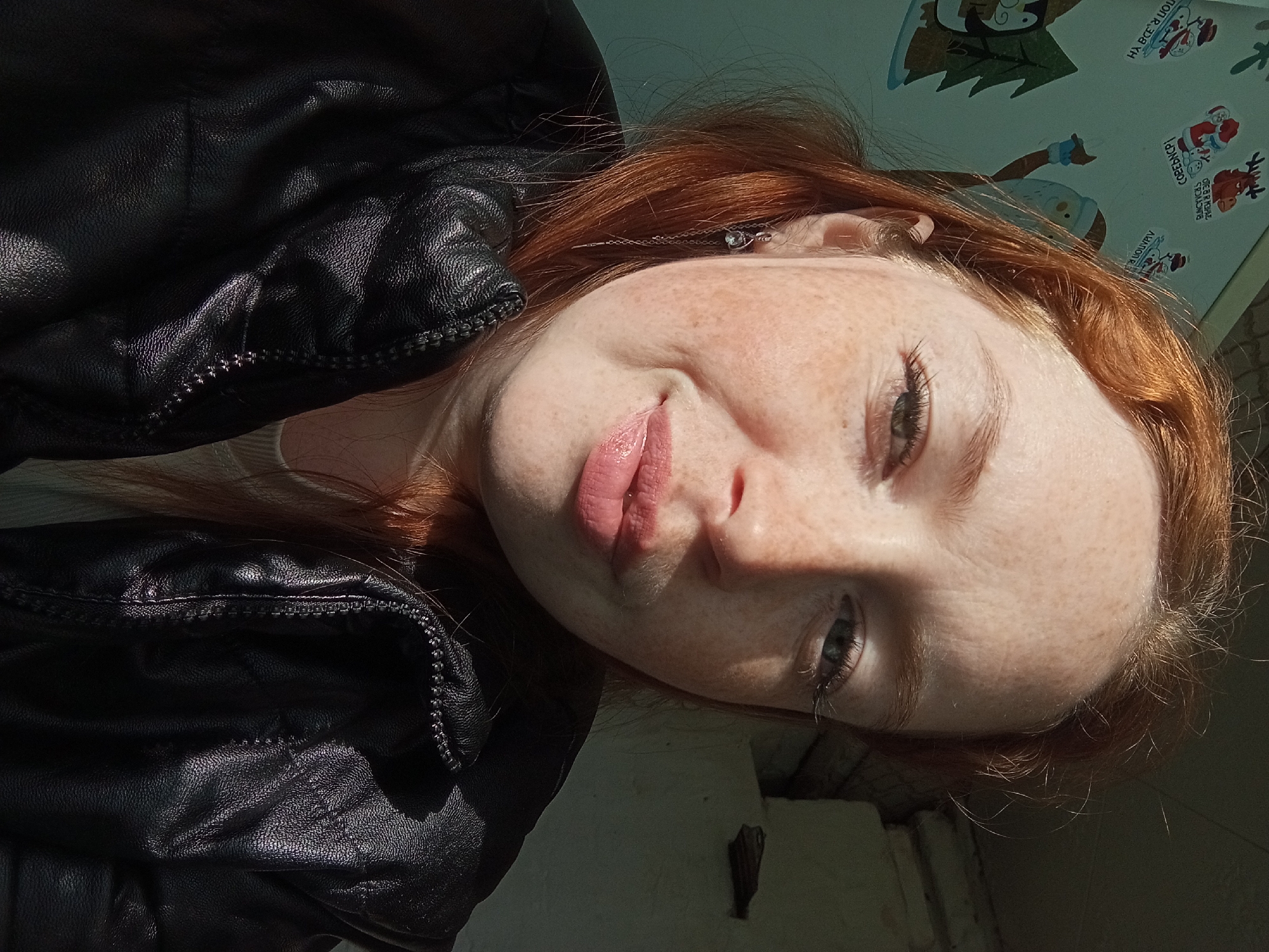 The image is a close-up selfie of a fair-skinned, red-haired woman with freckles, wearing a black leather jacket. The image has been rotated so the woman is tilted to the side. She is looking at the camera with a slight smile. The background is a wall with a sticker of cartoon Christmas figures. There is a partially visible white structure in the lower right. The location is identified as Yurgamysh, Russia.