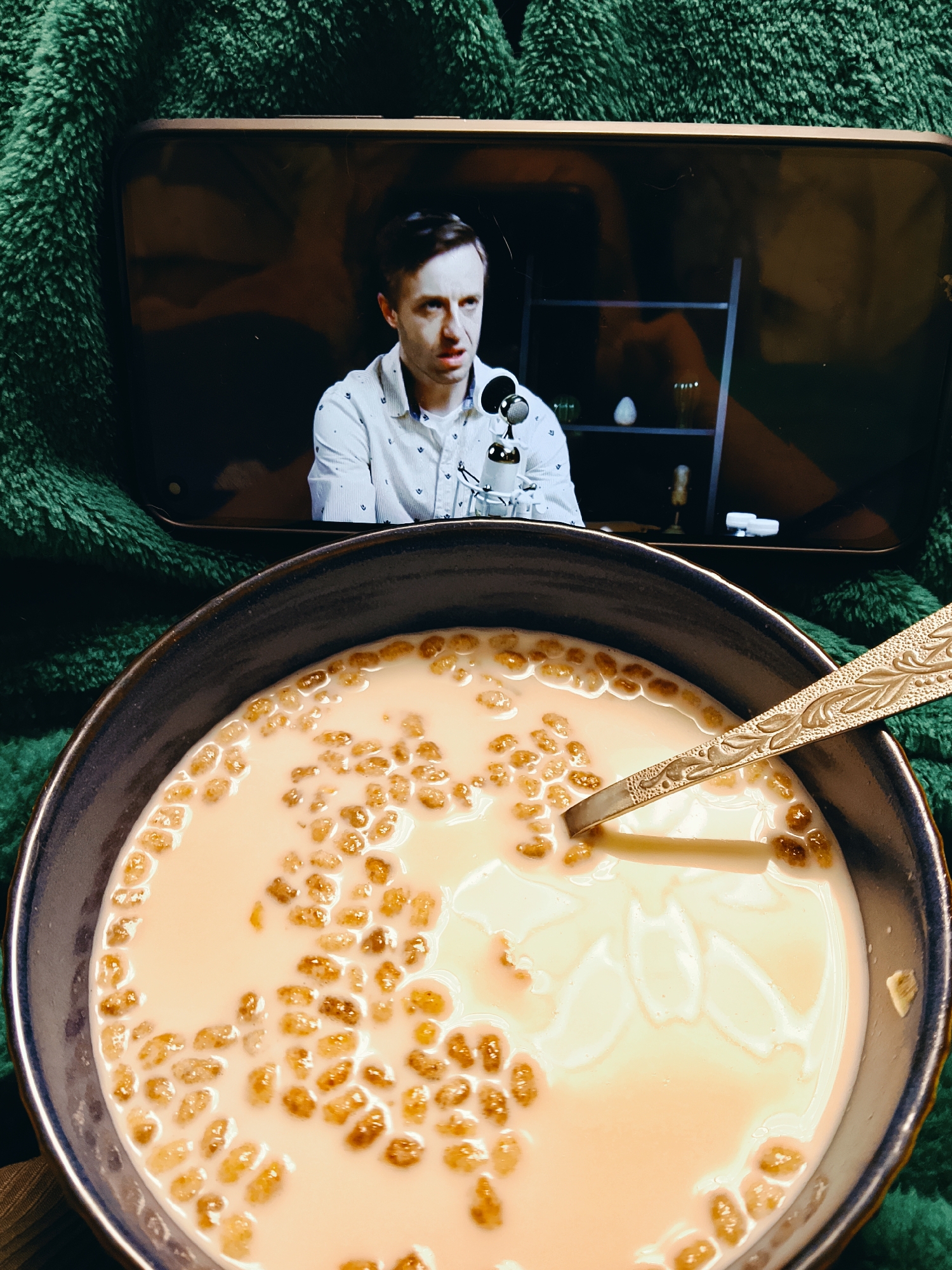 A photograph taken indoors shows a bowl of cereal with milk and a spoon, with a mobile phone displaying a man speaking into a microphone in the background. The cereal appears to be puffed rice. The bowl is dark with a blue rim and rests on a wooden surface and a green, fluffy blanket. The mobile phone displays a man in a light-colored patterned shirt talking, with a microphone in front of him and a bookshelf behind him. The spoon is metal with an ornate design. The environment suggests a casual, home setting. There is no indication that this was taken in Chaikovsky, Russia.