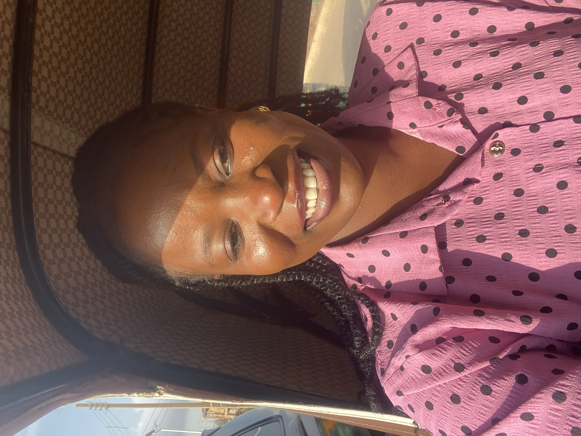 A young Black woman, likely in her early twenties, is captured in a selfie taken from inside a motorized rickshaw. She has a dark complexion and is wearing a pink collared shirt with black polka dots. Her hair is braided and extends down her back. The interior of the rickshaw appears to be lined with a patterned fabric. Sunlight casts a strong shadow across her face, highlighting her cheekbones and nose. The background suggests an outdoor urban setting, with hints of buildings and power lines visible through the open side of the rickshaw, indicating a location in Bukuru, Nigeria.