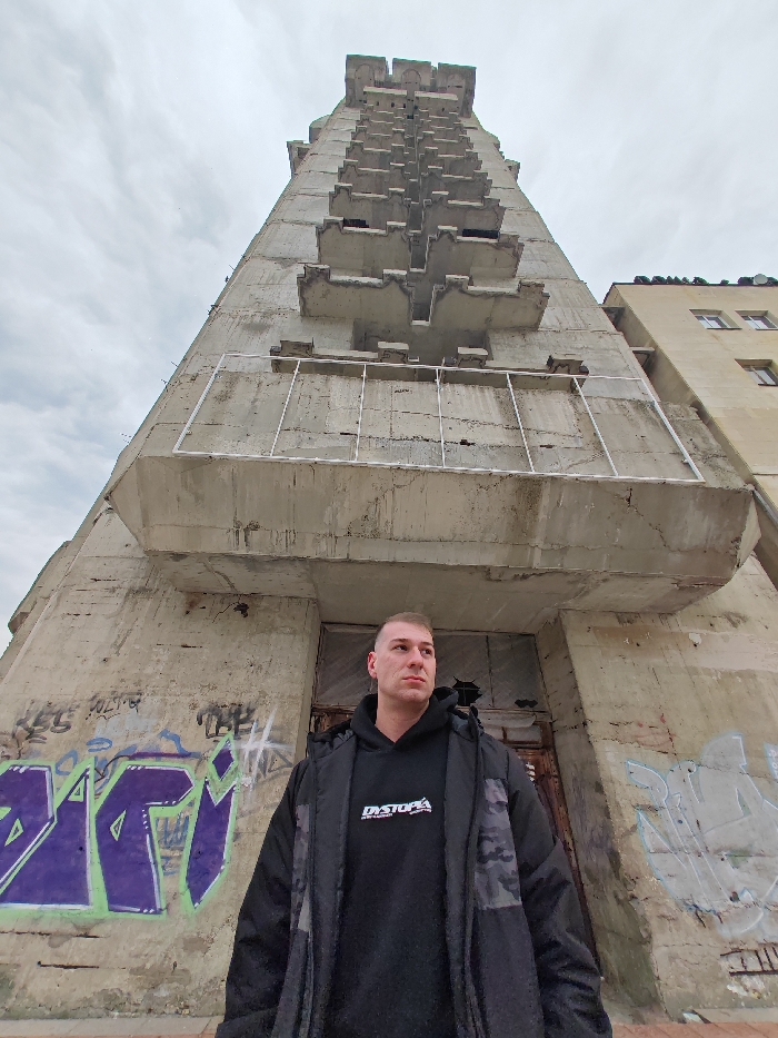 A low-angle photograph captures a male subject in Shumen, Bulgaria, standing before a prominent, multi-story concrete structure. The subject, a Caucasian male with short light-brown hair, is positioned in the lower-center right, wearing a black hooded sweatshirt with "DYSTOPIA" printed in white on the chest and a dark-colored parka featuring camouflage patterns on the shoulders and arms. He maintains a neutral expression, looking to his right. Behind him rises a tall, Brutalist-style concrete building, identifiable as the Unfinished Clock Tower of Shumen, which extends upwards under an overcast sky. The tower's facade is characterized by numerous vertically stacked, cantilevered concrete balconies or platforms that progressively decrease in size towards the apex. Many of these balconies lack complete railings; some exhibit partial white metal railing frames. The lower sections of the concrete walls display extensive wear, staining, and prominent graffiti, including large purple and green lettering on the left side. A dark, recessed entryway, appearing damaged or boarded up, is visible directly behind the subject. To the right of the concrete tower, a portion of a lighter-colored, multi-story building with windows is partially visible.