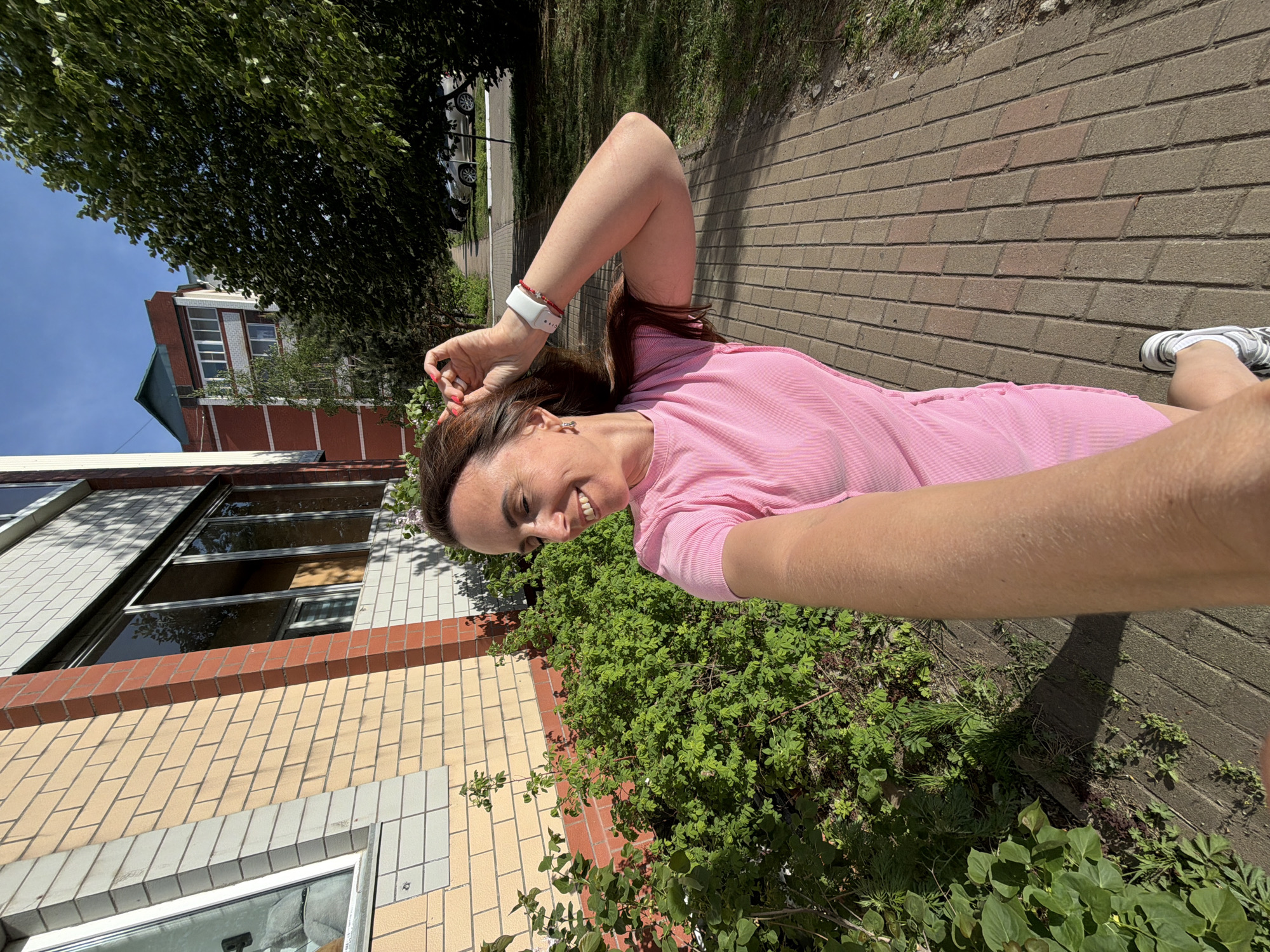 The image is a selfie taken outdoors. A woman with brown hair, wearing a pink shirt and a white fitness tracker on her wrist, is smiling at the camera. She's standing near a brick building and some greenery. Part of a street or sidewalk made of brick is also visible in the background, with a tree and possibly cars further away. It is daytime and the weather appears sunny.
This image does not contain adequate metadata to place this image at Birobidzhan, Russia.