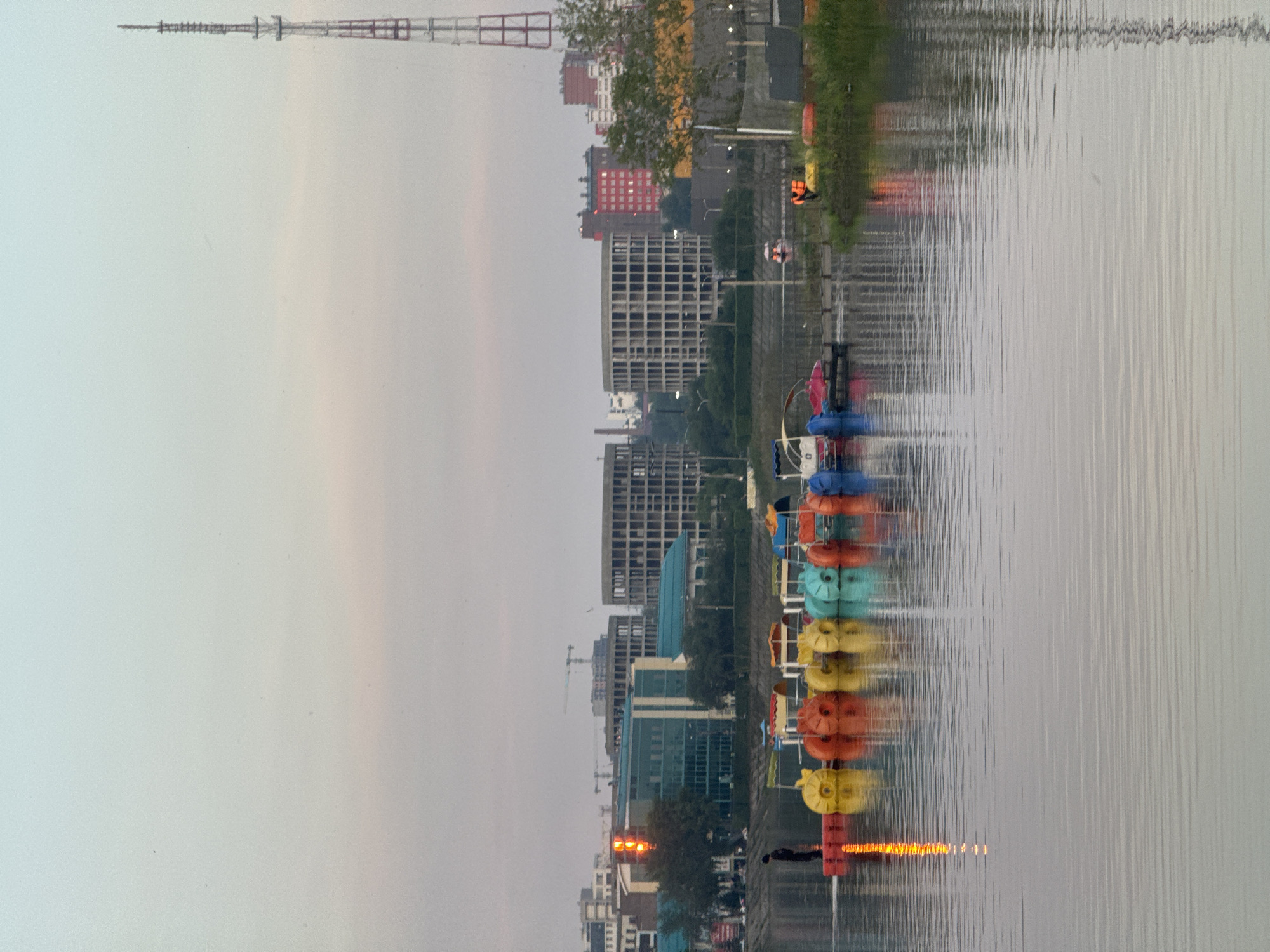 The image depicts a serene waterscape in Irkutsk, Russia, likely during twilight or early morning given the soft, diffused lighting and pastel hues in the sky. In the foreground, the calm water of a river or lake reflects the surrounding environment. A row of colorful pedal boats – yellow, orange, green, and blue – are moored to a red floating dock on the left side of the frame. One person stands on this dock, facing away from the camera, towards the water. In the middle ground, a line of buildings forms the cityscape. These include modern structures with glass facades and older, unfinished concrete buildings under construction. A prominent red and white radio tower stands tall on the right side of the image, a distinctive landmark. The overall atmosphere is tranquil, with a hint of urban activity in the background.