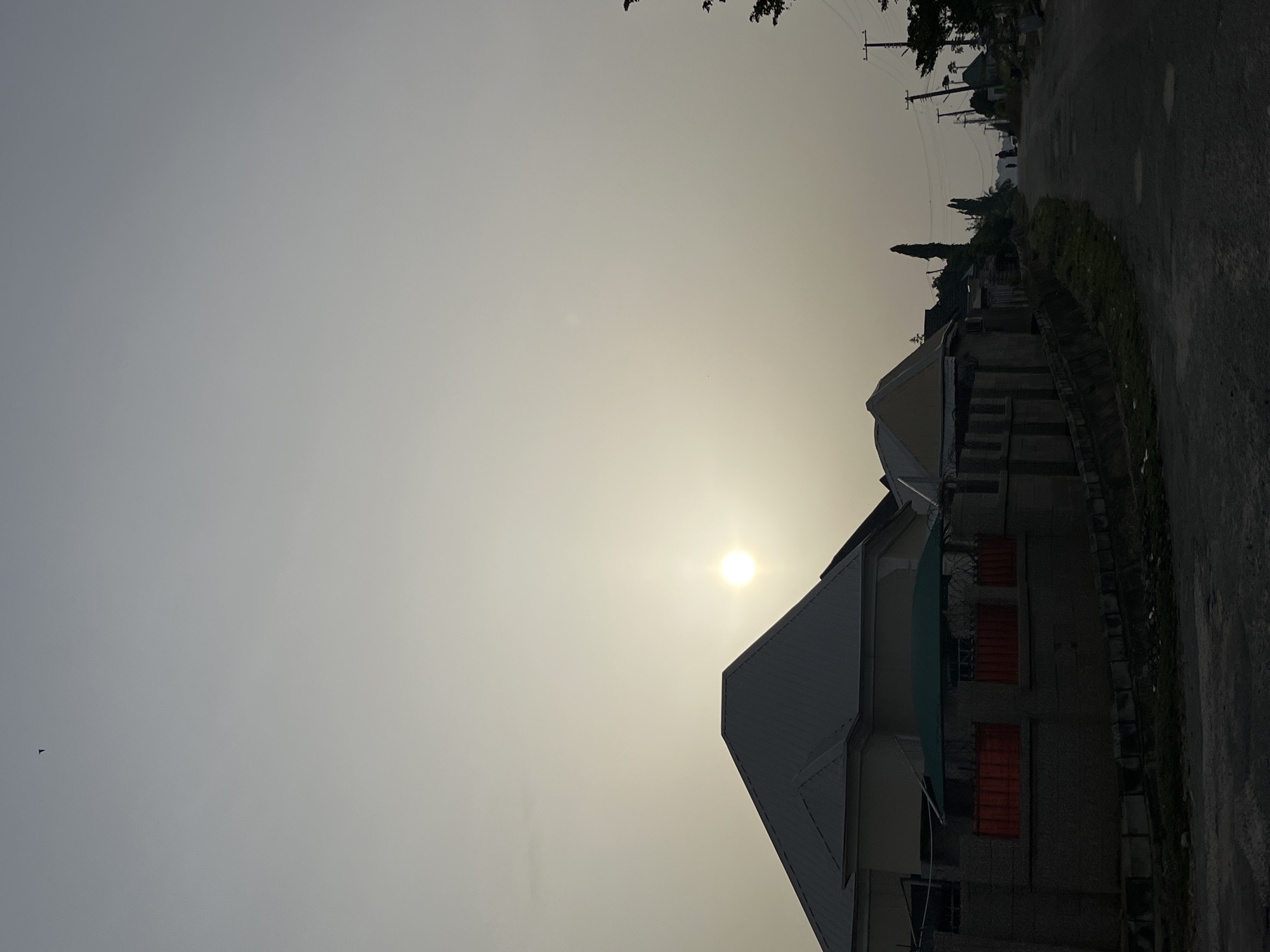 A view of the sky shows a bright, hazy sun positioned over a residential building in Abuja, Nigeria. The building has a sloped roof with at least two stories. Windows with red grilles are visible. The street runs along the side of the building, with utility poles visible further down the road. The sky is light grey.