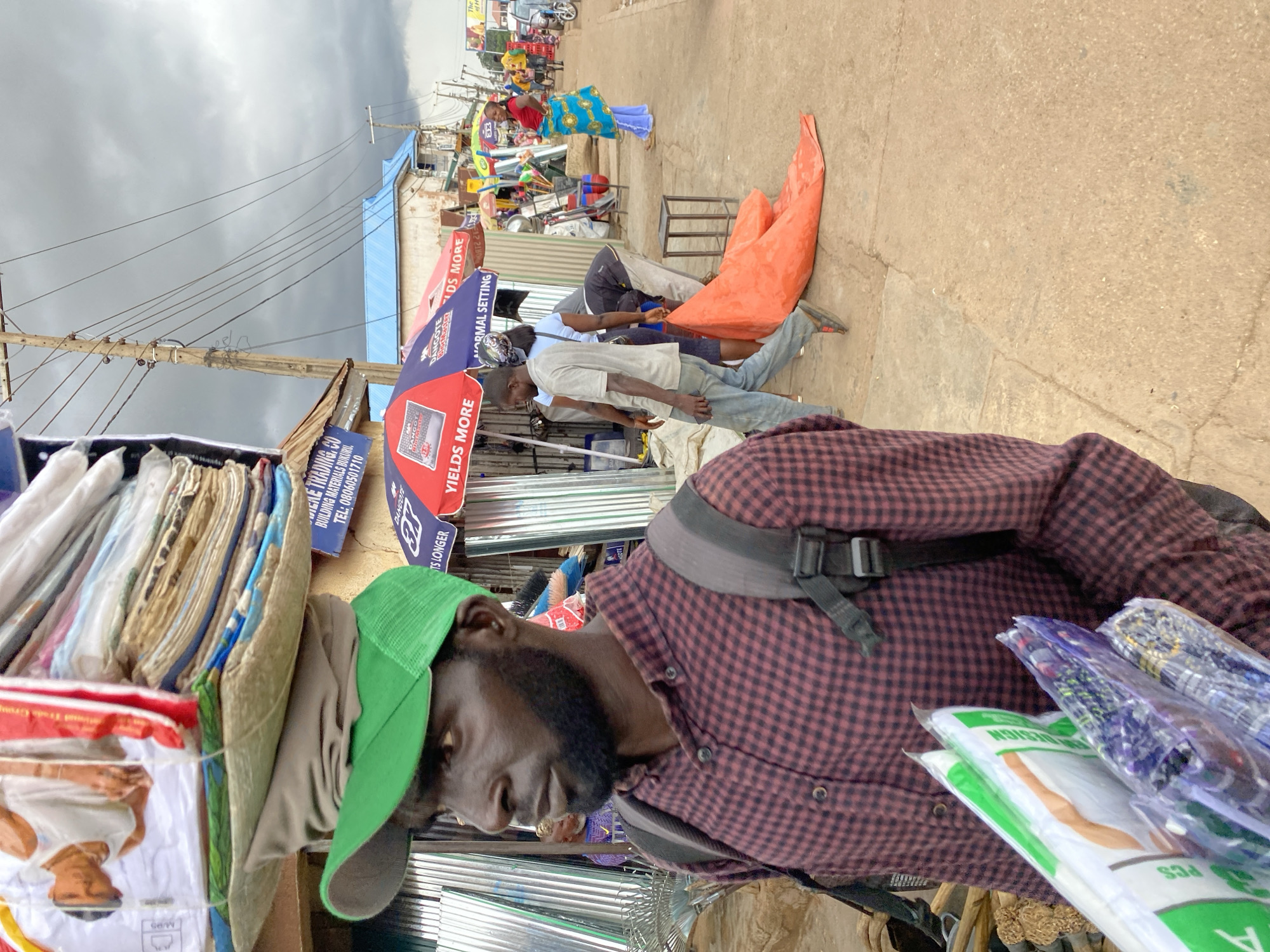 In Bukuru, Nigeria, a Black man with a beard and a green baseball cap carries a large, stacked load of merchandise on his head, including textiles. He wears a maroon and black checkered button-down shirt and a grey backpack. In front of him, he holds several packages, some with green and white labeling. The man is positioned on a dirt and concrete pathway, with a corrugated metal structure to his left. To his right, a street scene unfolds with other individuals and various market stalls. Umbrellas are visible, advertising "Dangote" and "YIELDS MORE." An orange tarp lies on the ground near a few individuals, one of whom is a woman in a blue and gold patterned skirt. In the background, buildings with blue roofs and utility poles with a dense network of wires are present under a cloudy sky. The overall environment suggests an outdoor market setting.