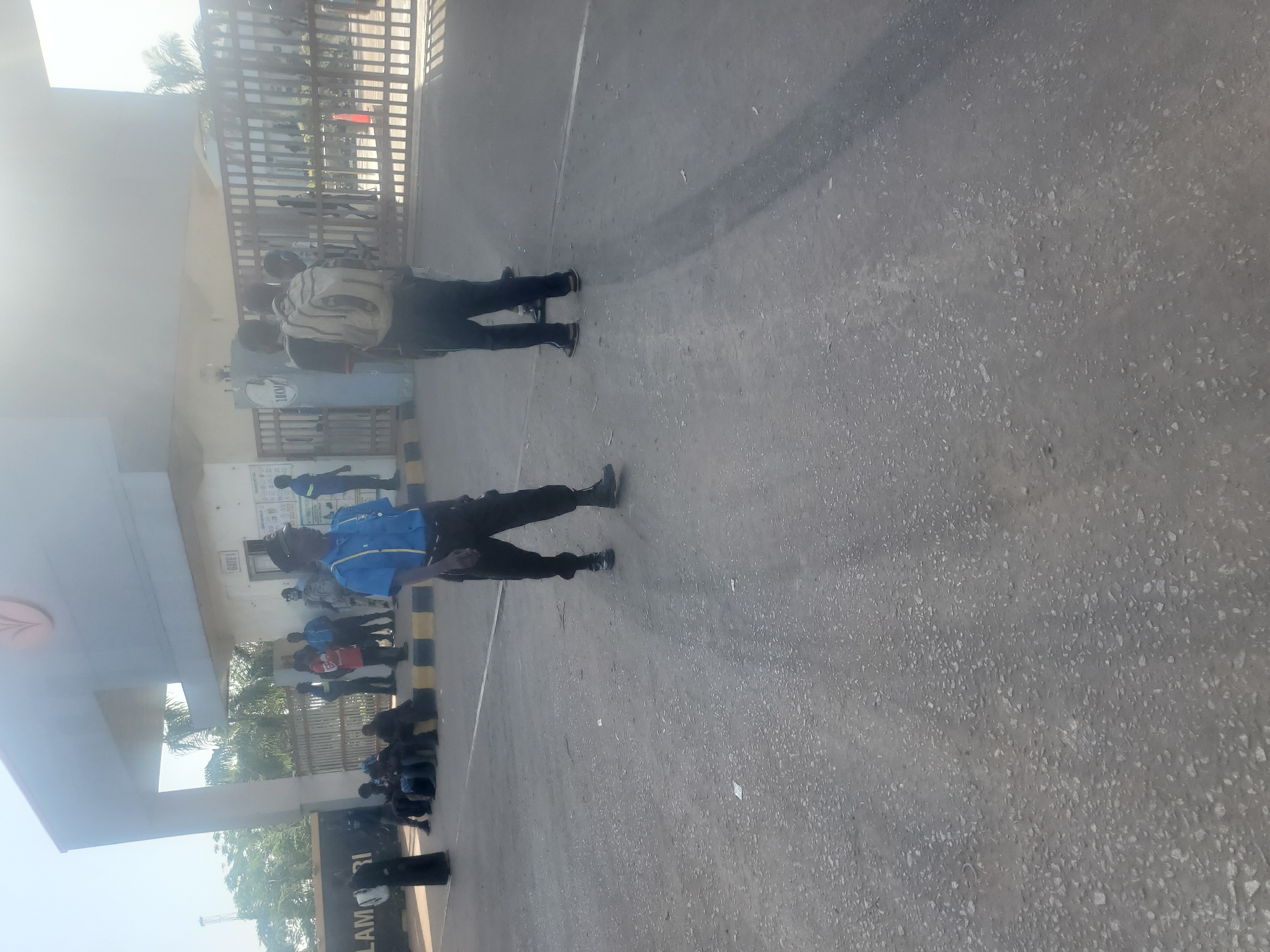 This image, taken in Gwagwada, Nigeria, captures an outdoor scene at what appears to be an entrance or gate to a facility, under bright daylight conditions.
In the foreground and midground, two individuals are standing on a grey asphalt surface. The person closer to the viewer is wearing a blue and yellow collared shirt with dark trousers, and black shoes. Slightly behind and to their left, another individual wears a light-colored, possibly striped, long-sleeved shirt, dark trousers, and carries a backpack.
To the left side of the frame, a group of people is gathered under a light-colored covered structure, which appears to be a gatehouse or shelter. This structure features a pillar with a stylized leaf or tree logo. On the wall of the structure, a white sign clearly indicates "GATE I," and another board displays some indistinct text and diagrams. Black and yellow striped bollards line the edge of the asphalt surface, marking a boundary.
Further back, behind the covered structure and bollards, a large metal gate is visible. Several more individuals are gathered near and behind this gate, with some standing and others appearing to be sitting or crouching on the ground. A partial sign on the ground or a low wall in the lower left corner of the frame (when correctly oriented) shows the text "LAM RI." The ground is an asphalt surface with scattered light-colored debris. The bright sunlight suggests it is daytime, casting subtle shadows and creating a slight glare in the upper left part of the image. The scene depicts people congregating at a secured entry point.