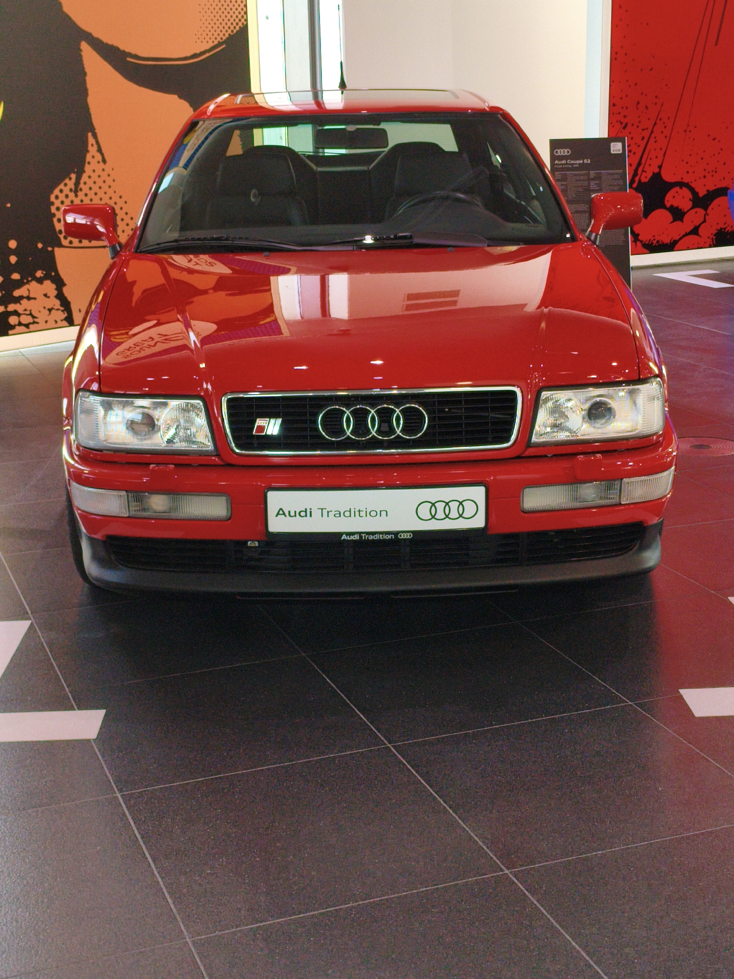 A red Audi Coupé S2 from 1990 is displayed indoors. The car faces forward, centered in the frame, with its headlights illuminated. A license plate on the front reads "Audi Tradition." The car is positioned on a dark tiled floor with white lines. In the background, a large orange abstract design is visible on the left, and a red graphic design is on the right. A white column is present in the far background. A placard displaying "Audi Coupé S2" and other information is mounted to the right of the car. The scene appears to be a showroom or exhibition space, likely within the Audi Forum in Ingolstadt, Germany. No people are visible in the image.