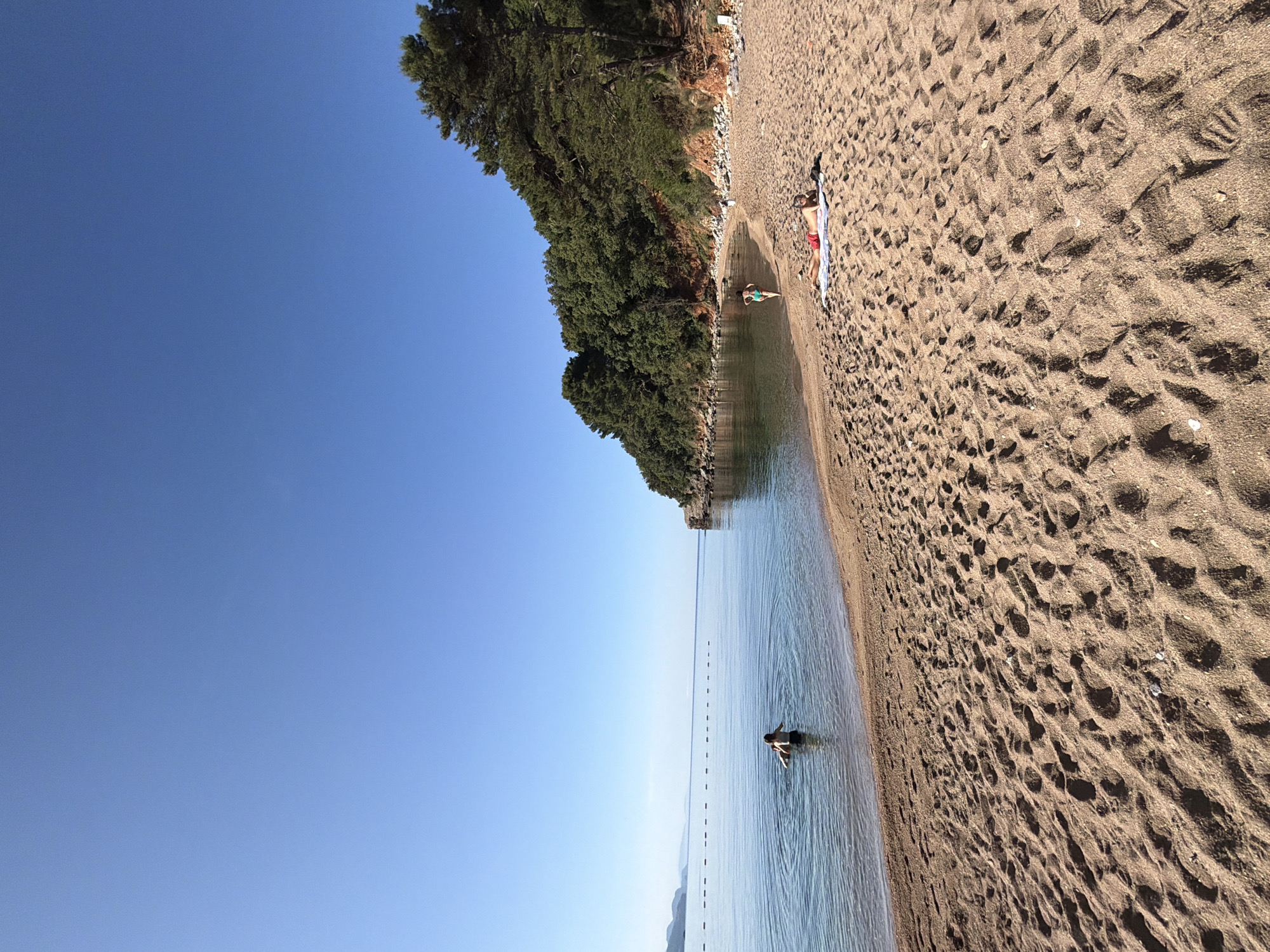 The image depicts a tranquil coastal scene on a sunny day at a beach in Budva, Montenegro. In the foreground, a sandy beach extends across the lower portion of the frame, characterized by numerous footprints and a golden-brown hue. One individual, wearing red swim trunks, lies prone on a white towel, sunbathing in the mid-right. A second individual, wearing a teal bikini, walks from the water onto the beach near the base of the forested hill on the right.
The midground features calm, clear blue water of a bay. A third individual, wearing dark swim trunks, stands waist-deep in the water on the left, facing the distant horizon, with visible ripples emanating around them. A series of white buoys marks a boundary across the water, extending horizontally from the left side of the frame towards the right. The right side of the bay is framed by a steep, rocky hill densely covered with vibrant green trees, predominantly pines.
In the background, the serene sea meets a clear, cloudless, deep blue sky at the horizon. Faint outlines of distant landmasses or mountains are visible on the far left, suggesting a continuation of the coastline. The scene is illuminated by bright daylight, casting minimal shadows and indicating fair weather.