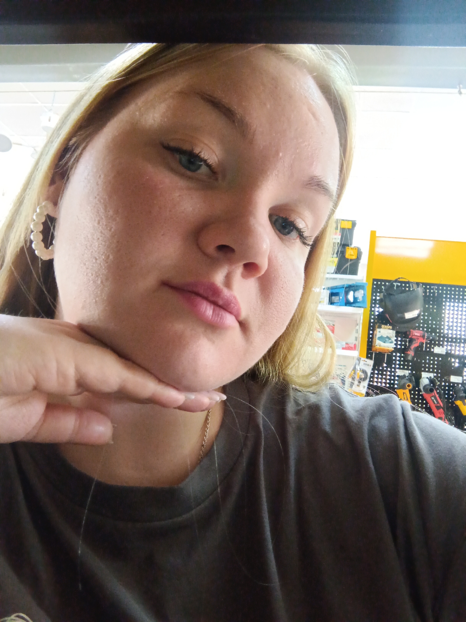 A fair-skinned young woman with blond hair is taking a selfie. Her left hand is posed beneath her chin. She wears pearl earrings, a light-colored necklace, and a grey t-shirt. The background appears to be a retail environment with shelves displaying various items, some of which are tools or related equipment. The colors yellow, black, and blue are prominent in the background. The location is most likely in Фоки, Russia, based on user request.