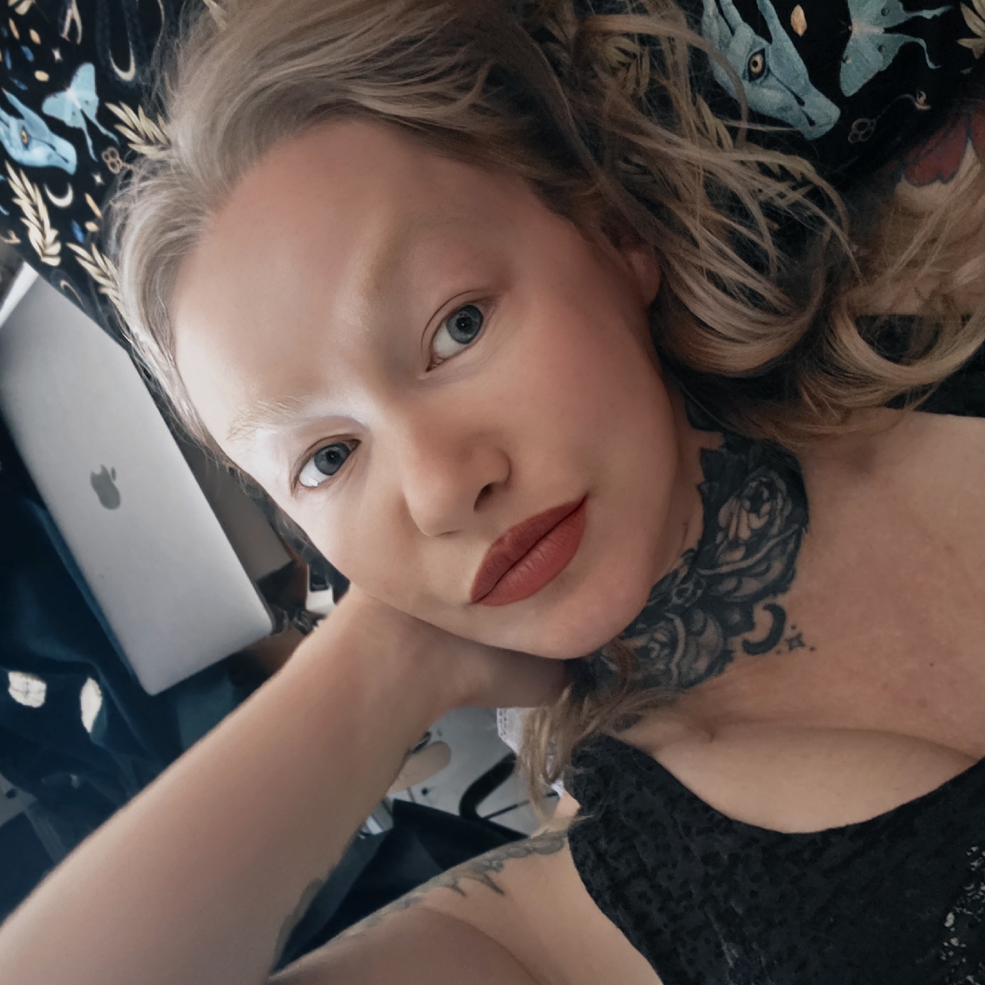 A high-angle, close-up selfie depicts a fair-skinned person with blue eyes and light blonde hair, partially obscured by the framing. The person's eyebrows appear bleached or very light. They are wearing a dark-colored top with a textured pattern. A prominent tattoo of a floral design is visible on the neck and upper chest, with additional tattoos on the arm. The person rests their head on a light-skinned arm and hand. An open Apple laptop is partially visible in the background. The background also features a dark fabric with a design of blue dragons and moons. There is no indication of Saint Petersburg, Russia, in the image.