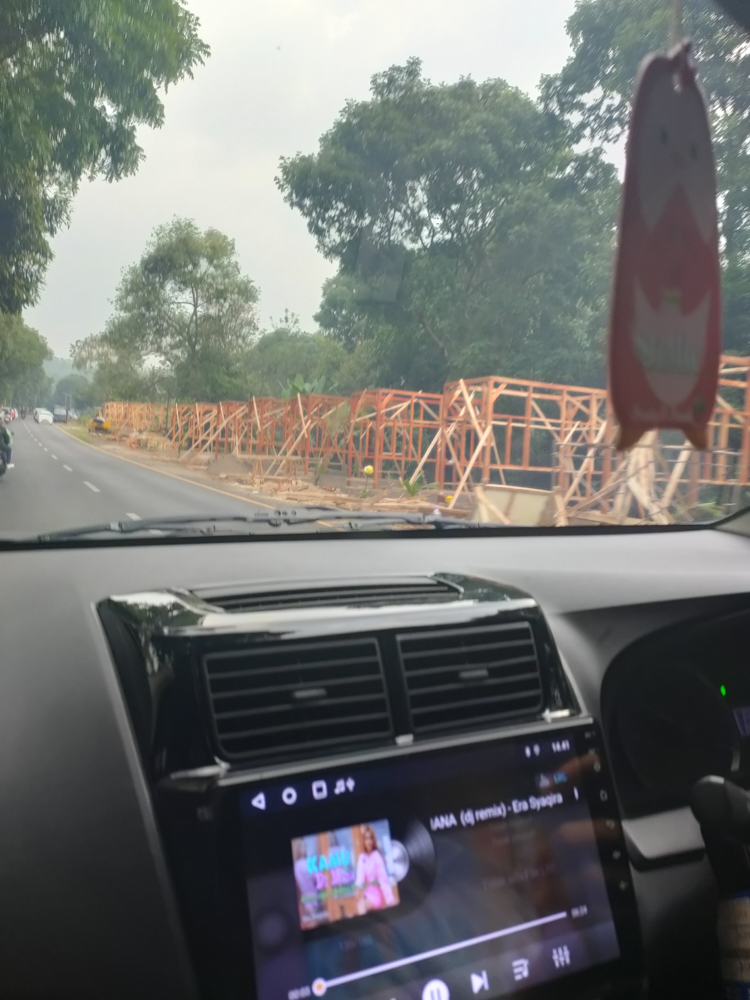 The image shows a view from inside a car driving on a road in Subang, Indonesia. The car's dashboard is visible, with a touchscreen infotainment system displaying song information. Outside the car, the road is lined with trees and a construction site where wooden frames are being erected. The sky is overcast. There are no discernible people or specific landmarks that would allow for further precise localization within Subang.