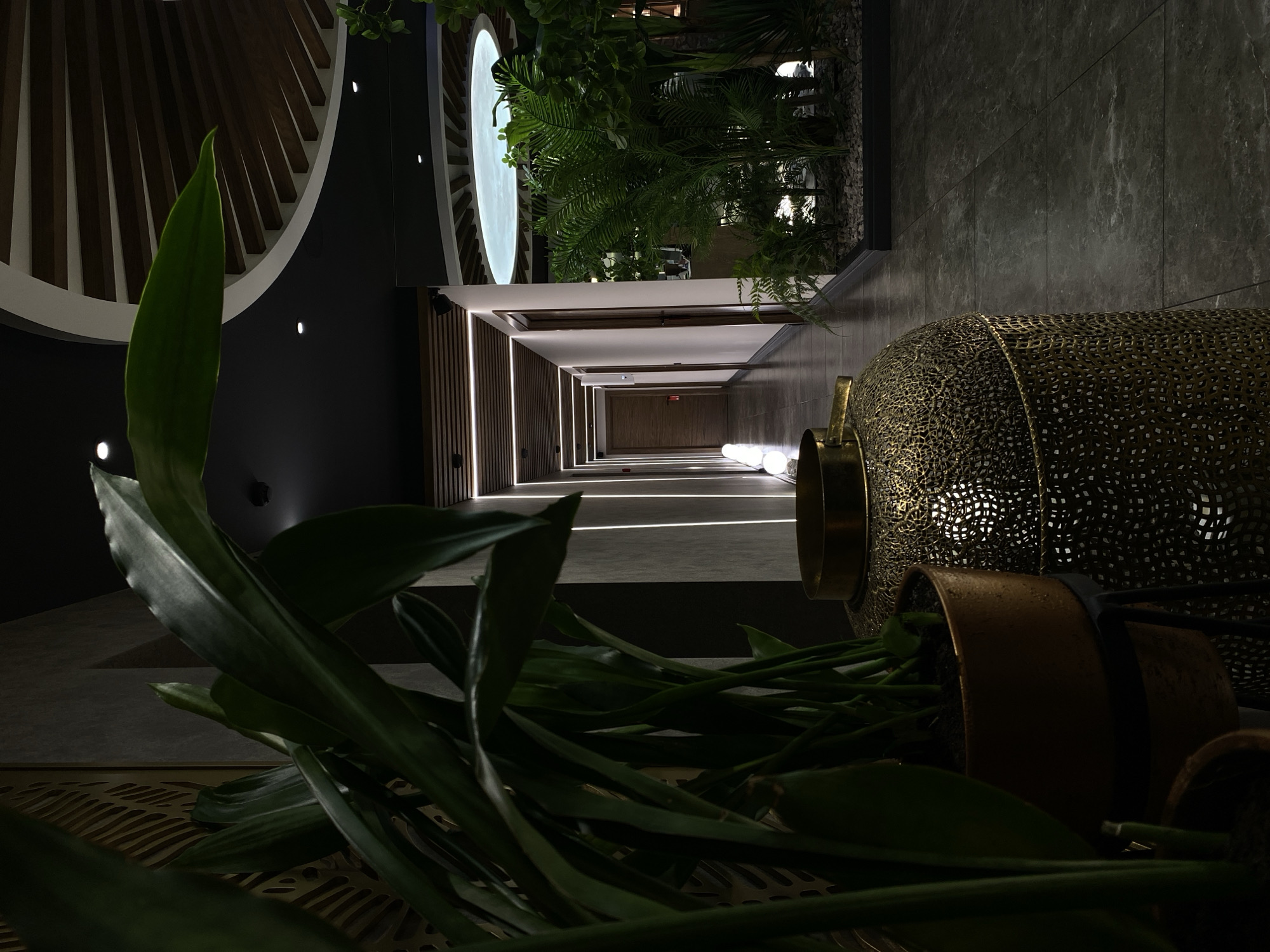 The image depicts an interior scene, possibly a hotel hallway or a contemporary building lobby, with lush greenery in the foreground and a long, well-lit corridor extending into the distance. The lighting is dramatic, with vertical lines of light accentuating the corridor walls, and a large, ornate golden lantern-like object is prominently featured on the left side, partially obscuring large green plant leaves. The corridor has multiple wooden doors on either side and is illuminated by recessed lighting and spherical light fixtures. The ceiling features an interesting architectural design with wooden beams and a circular recessed light fixture. The overall aesthetic suggests a modern and upscale environment. There are no people or specific Votkinsk landmarks visible.