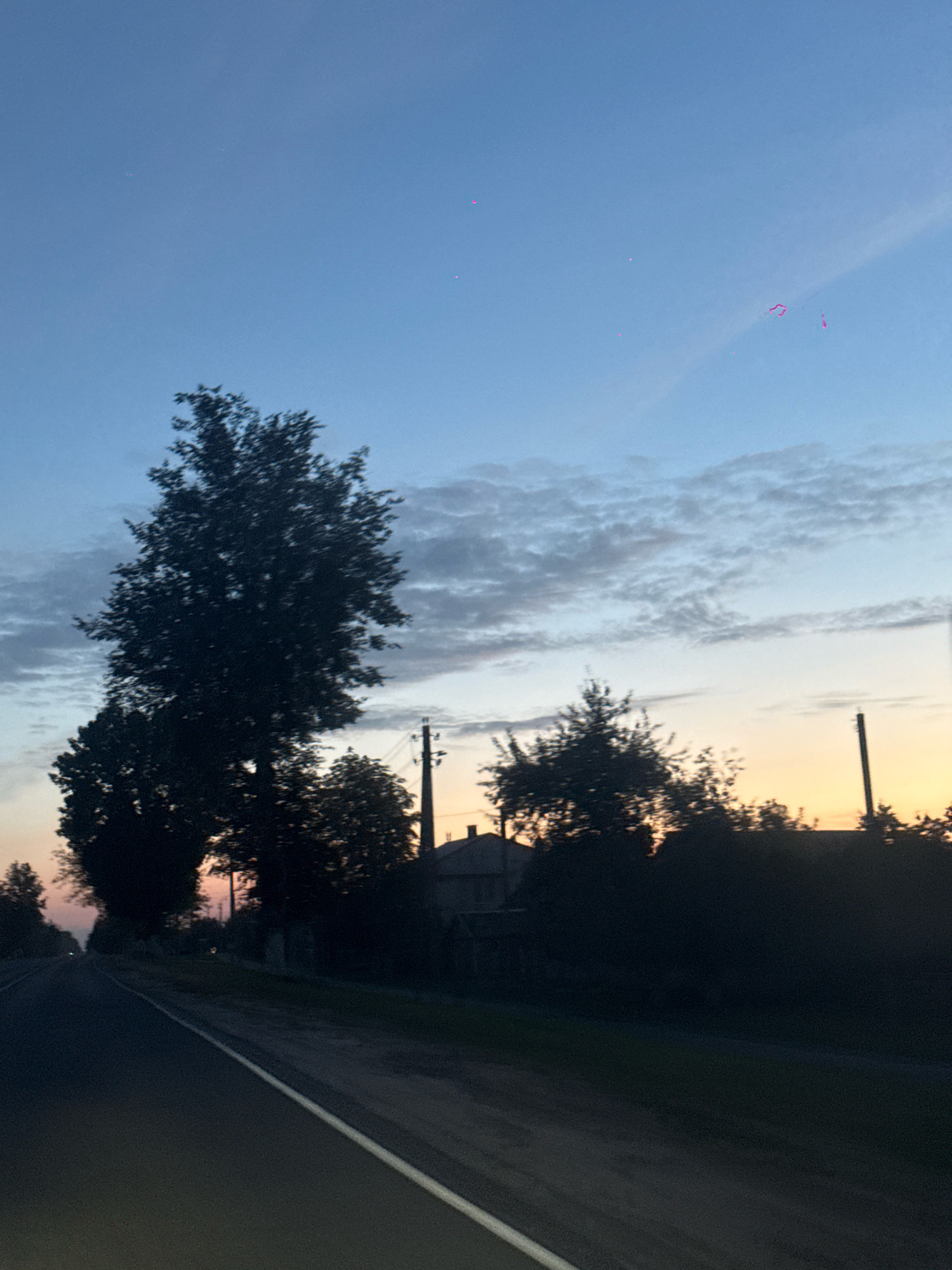 The image depicts a rural landscape in Slawharad, Belarus, at dusk. A paved road runs diagonally from the bottom left to the center of the frame, with a white line marking its edge. Silhouetted trees and a building with a visible power line are visible along the right side of the road. The sky is a gradient of deep blue at the top, transitioning to a pale orange at the horizon, with scattered clouds. Several small, indistinct pink or magenta lights are visible in the upper portion of the sky, possibly from a light pollution source or atmospheric phenomenon. The overall scene suggests a quiet, twilight atmosphere.