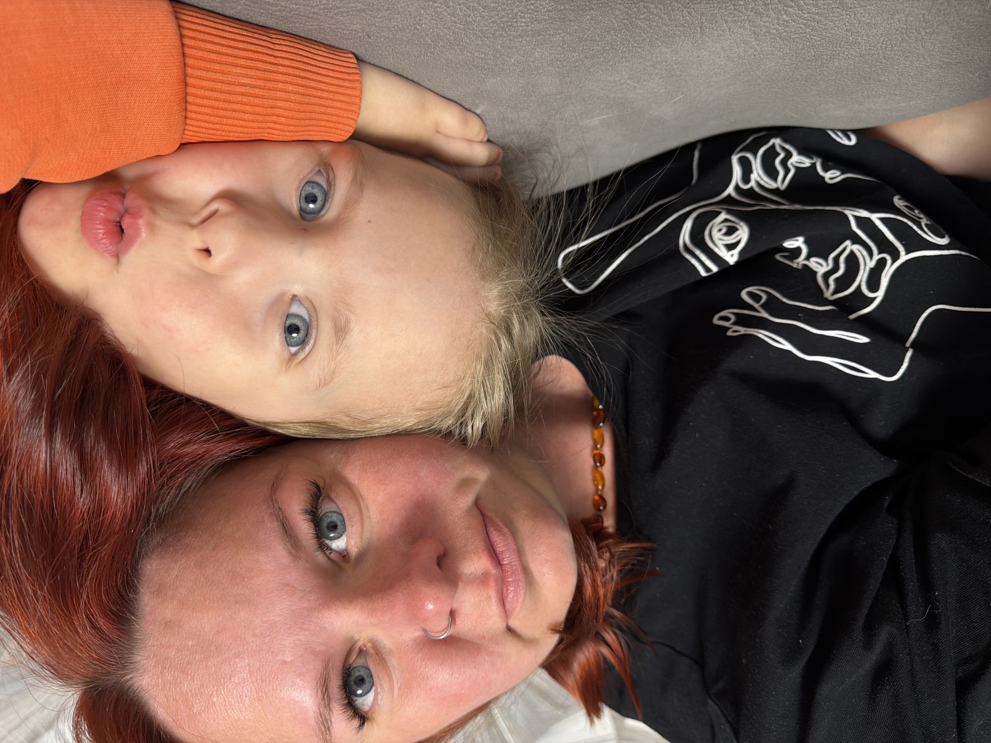 The image depicts two individuals lying on a soft surface. The lower individual is an adult woman with red hair and fair skin. She has a nose ring and is wearing a black shirt with white line drawings. The upper individual is a young child with blonde hair, fair skin, and blue eyes. The child is wearing an orange sweater. They are positioned head-to-head, with the child's head near the top of the image and the adult's head near the bottom. The background appears to be a gray or neutral-colored surface, possibly a couch or bed. There is no evidence of the location in Chaikovsky, Russia.