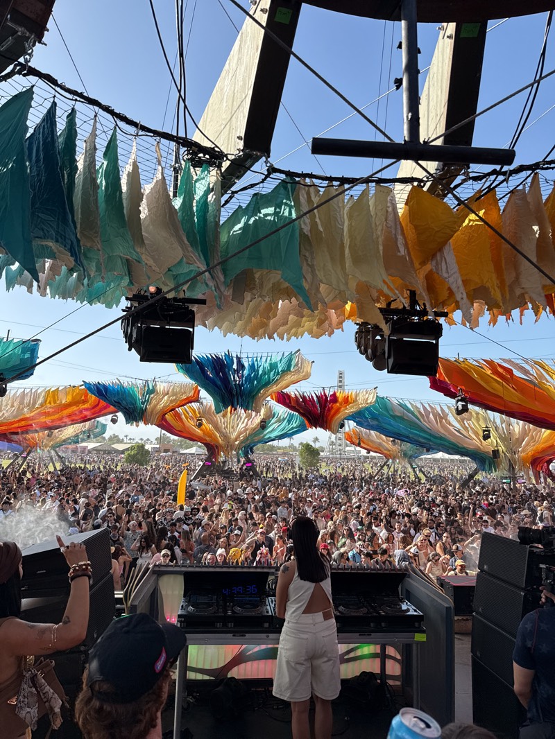 The image shows a crowded outdoor festival in Indio, California. A DJ, seen from behind, is operating equipment on a platform. A large crowd extends into the background, beneath colorful overhead installations resembling trees or flowers. Above the DJ platform, a circular structure supports hanging, multicolored decorations. Stage lights and sound equipment are visible around the DJ setup. A few people are near the stage. The weather appears clear and sunny.