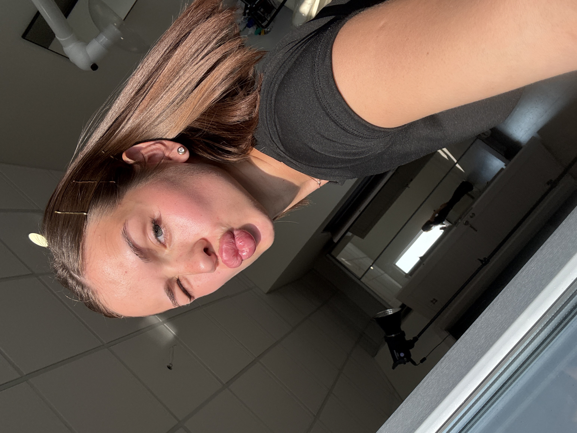 A young woman with medium brown hair and a black t-shirt poses for a selfie, puckering her lips and winking one eye. She is wearing a small stud earring in her visible ear and has several golden bobby pins in her hair. The lighting suggests natural sunlight coming from a window, casting a warm glow on her skin. The background appears to be an indoor setting with a tiled ceiling and a window with blinds. There are some objects visible in the background, including a light stand with a black reflector and some office-like furniture. The image is a close-up, focusing on the woman's face and upper body. The location is not identifiable as the city of Perm, Russia.