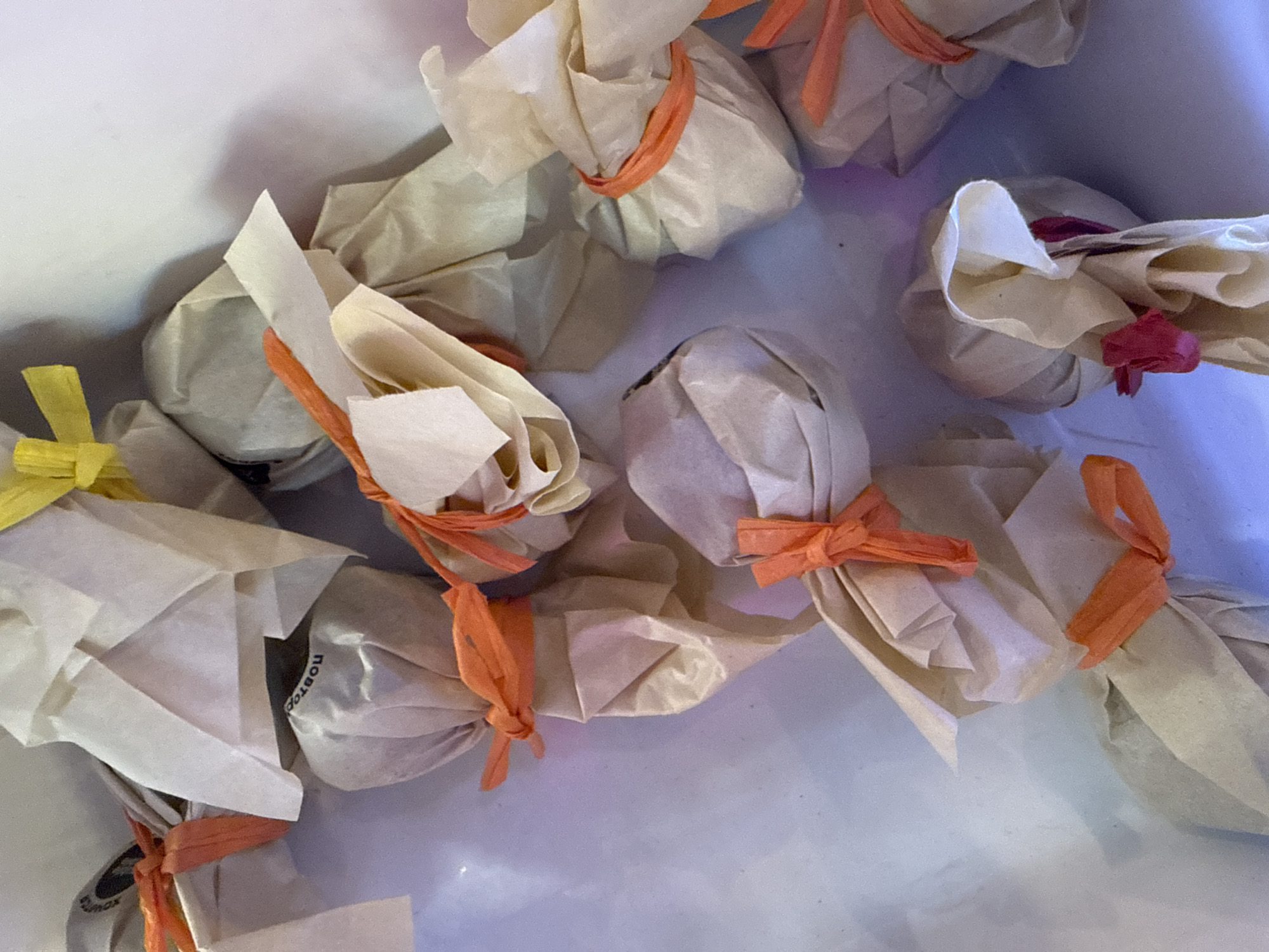 A close-up overhead shot shows a collection of individually wrapped items, likely gifts or treats, arranged on a white surface. Each item is encased in light brown and cream-colored paper, secured with colorful raffia ties in shades of orange, yellow, and pink. The paper is folded and twisted to create decorative bundles, some with multiple layers. The background is a soft, diffused white with hints of purple and blue light, suggesting artificial illumination. There are no people or specific Moscow landmarks visible in the image.