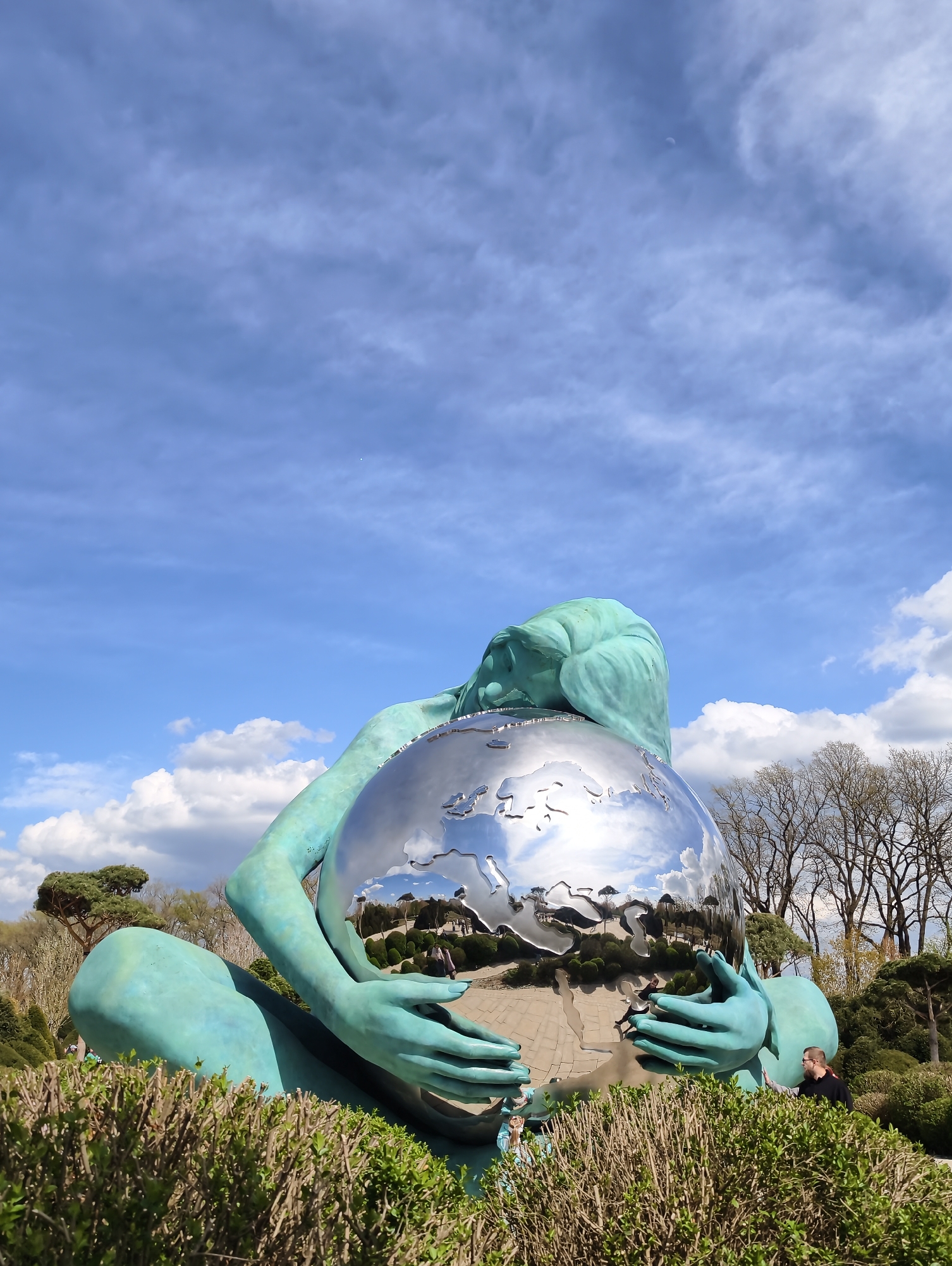 The image shows a large, turquoise-colored sculpture of a woman cradling a reflective, spherical globe. The globe reflects the surrounding environment, including trees, the sky, and people. The sculpture sits within a garden or park setting, with trimmed hedges visible in the foreground and trees in the background. The sky is blue with scattered white clouds. The location is likely Krasnodar, Russia. A person in a dark shirt is standing near the sculpture.