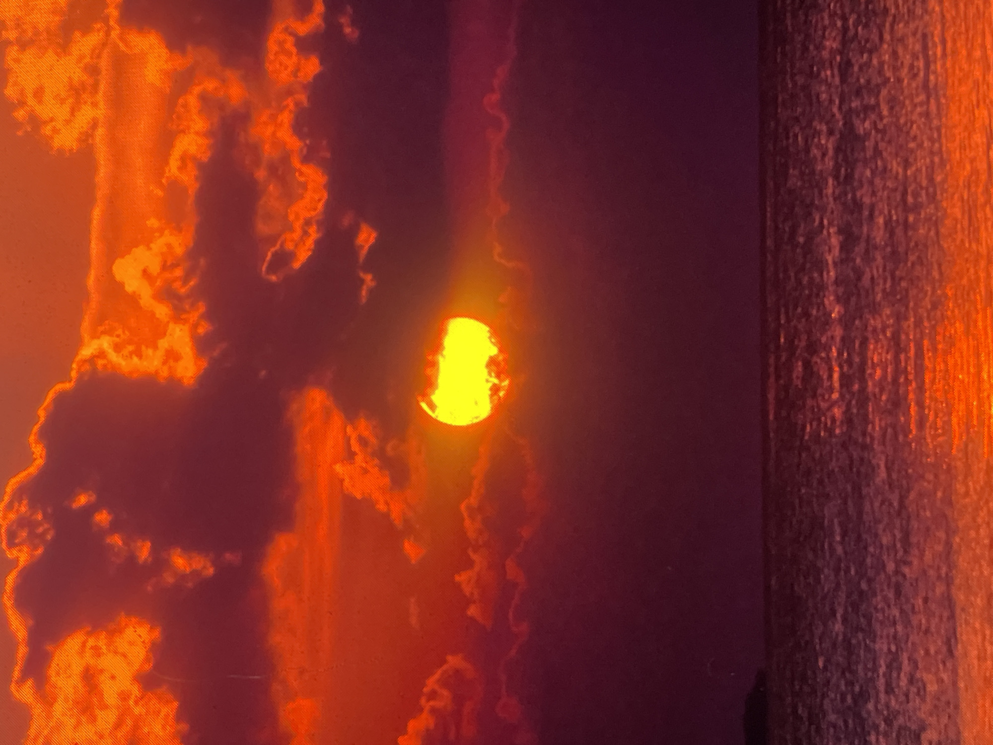 The image displays a vivid sunset or sunrise over a body of water. The sky, occupying the upper half of the frame, is dominated by intense hues of orange, deep red, and purple. Numerous clouds are present, some appearing as dark silhouettes against the bright backdrop, while others are illuminated with fiery orange and red edges, particularly in the upper and mid-portions of the sky.
The sun, a brilliant yellow-orange orb, is partially visible and appears to be setting or rising behind a dark cloud formation slightly left of the image's vertical center. Its light illuminates the surrounding clouds and casts strong colors across the scene.
The lower half of the image depicts a body of water, reflecting the dramatic colors of the sky in shades of deep red, purple, and orange. The surface of the water shows subtle ripples and reflections, particularly in the foreground. A distinct horizon line separates the sky and the water. On the far left, a small, dark, and indistinct landmass or distant coastline is barely visible against the water.
There are no discernible people, specific subjects (other than the natural elements), objects, or actions present in the image. The image exhibits visible digital artifacts, including pixelation and faint horizontal scan lines. The image provides no discernible geographical markers or specific landmarks to confirm its location as Zelenyi Hai, Ukraine.