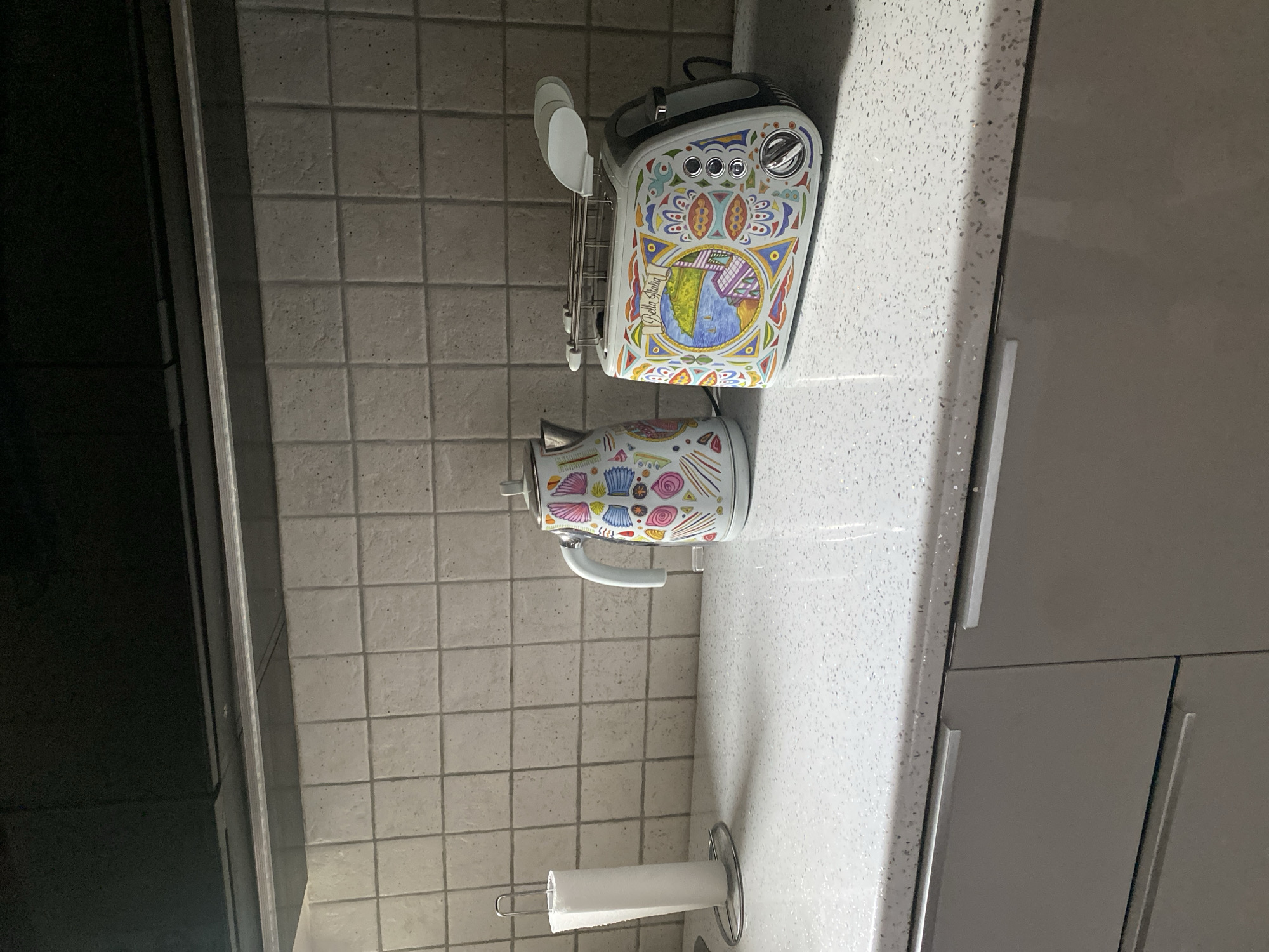 This is a medium shot, eye-level photograph of a kitchen counter with a tiled backsplash. On the white, speckled countertop, there is a white paper towel holder with a roll of white paper towels on the left. To the right of the paper towel holder is a white electric kettle with a light blue handle, decorated with colorful, abstract patterns. Next to the kettle is a white toaster with a similar colorful, decorative design, featuring a landscape scene with the text "Bella Italia". The toaster has chrome accents and a silver dial. The kitchen cabinets below the counter are a matte grey color with horizontal silver handles. The backsplash is made of small, square, light grey tiles with darker grout lines. The lighting in the image is dim, casting shadows from the objects onto the countertop. The background above the backsplash appears to be a dark brown or black cabinet or fixture. The scene does not provide any information about the city of Wrocław, Poland.