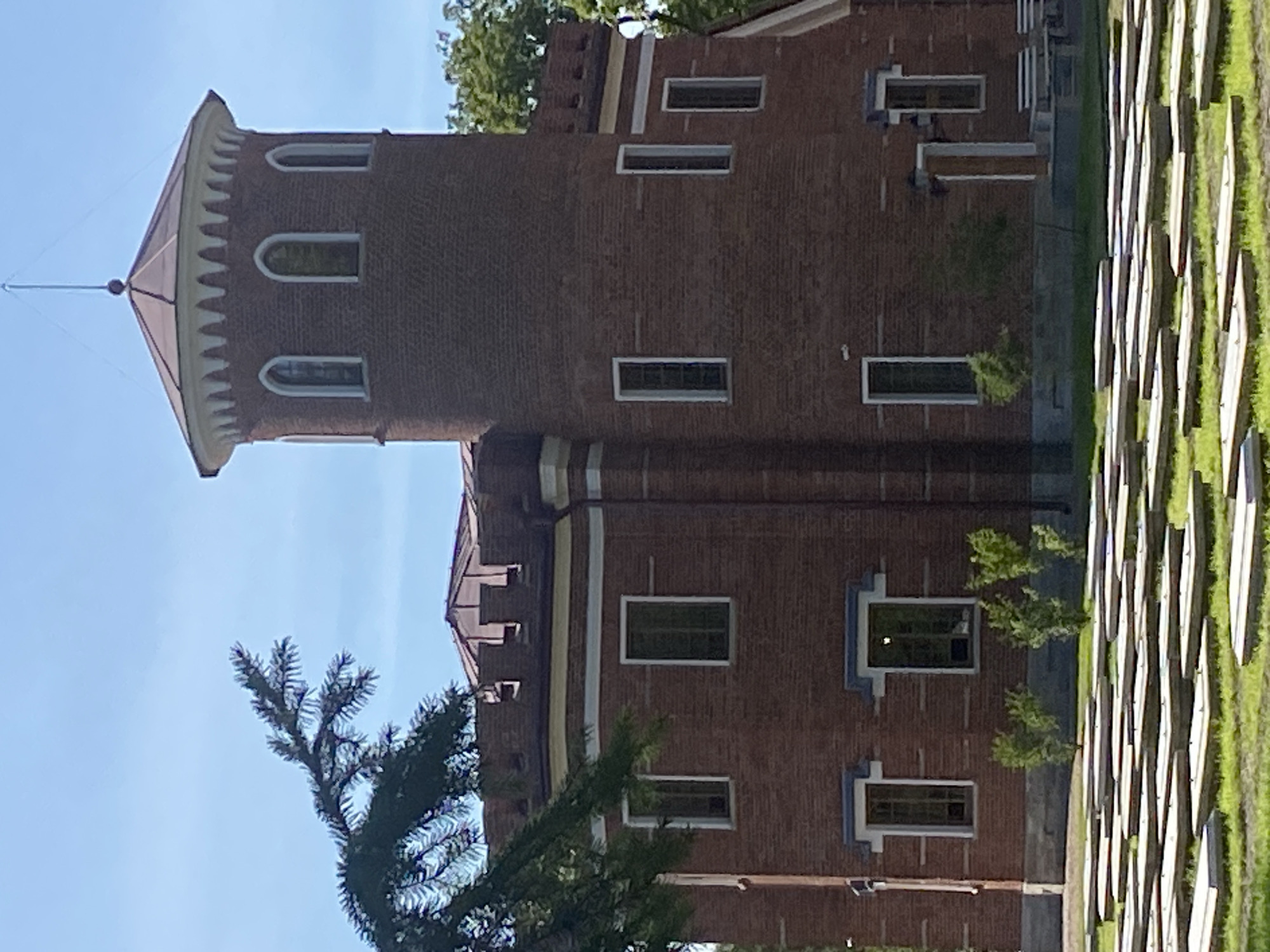 The image depicts a red brick building with a cylindrical tower, reminiscent of a castle, located in Pushkin, Russia. The building features a crenellated parapet along its upper edges and arched windows on the tower. In front of the building, a grassy area is laid out with rows of rectangular, light-colored objects, possibly benches or seating, arranged in a grid pattern. A dark green coniferous tree branch partially obscures the upper left portion of the image. The sky is clear and blue.