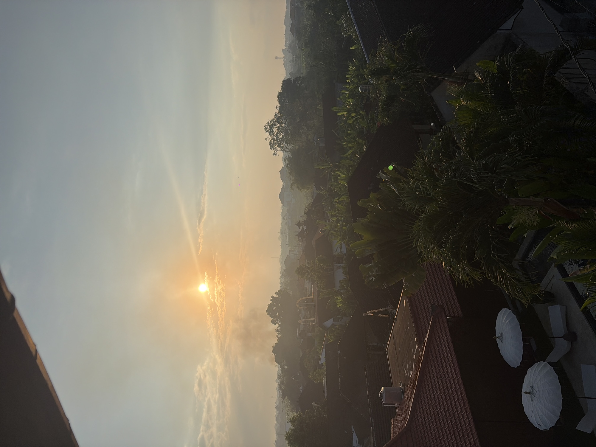 The image captures a sunrise over a residential area in North Kuta, Indonesia. The sun, a bright orb, is positioned in the upper left quadrant of the sky, casting a warm, golden light. Wisps of smoke are visible in the distance, rising from behind a line of trees. The foreground features several rooftops, some with red tiles, interspersed with lush green foliage. In the lower left, two white umbrellas are set up over lounge chairs next to a swimming pool, suggesting a resort or private residence. The scene is hazy, typical of a warm, humid morning in a tropical climate.
