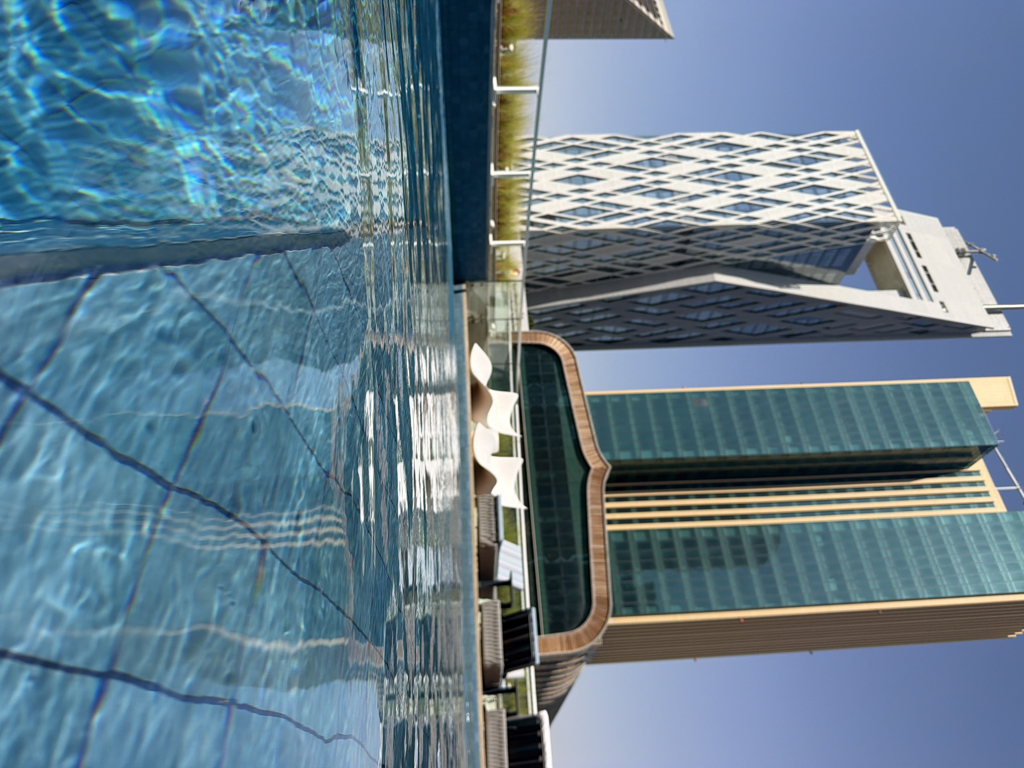 This image depicts a low-angle view of a swimming pool with a city skyline in the background. The pool water is clear blue, with reflections of the sky and buildings. In the background, there are two prominent skyscrapers, one with a distinctive white geometric facade and the other a tall, rectangular building with a teal glass exterior. In front of the skyscrapers, there are lounge chairs and some greenery, suggesting a rooftop setting. The sky is a clear, bright blue. The image appears to have been taken on a sunny day.