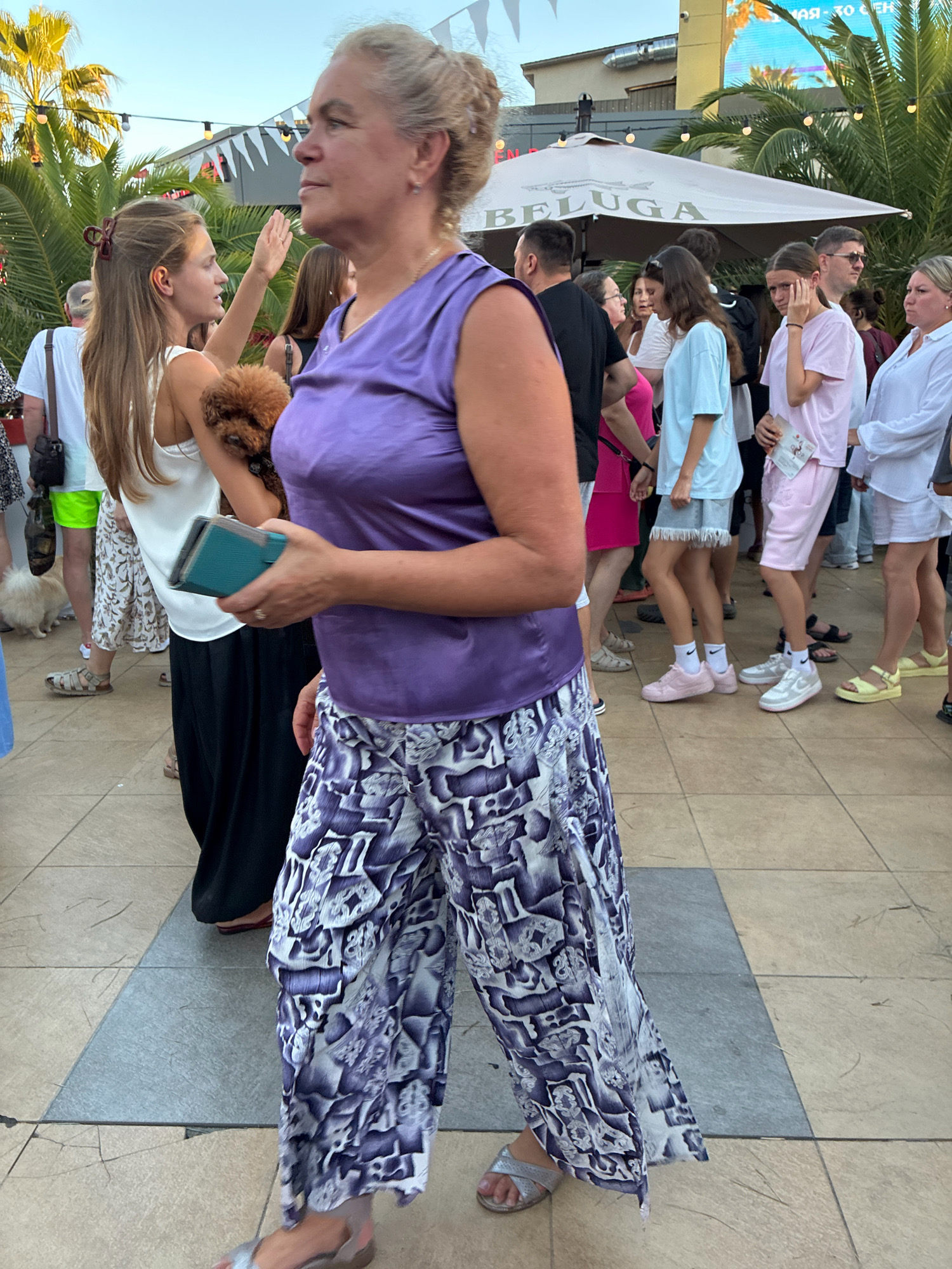 The image depicts an outdoor scene in Sochi, Russia, likely during the day or early evening given the lighting. A group of people are gathered, some walking and others standing. In the foreground, a woman with blonde hair pulled back is wearing a sleeveless purple top and wide-leg pants with a white and purple abstract pattern. She is holding a dark turquoise object, possibly a phone or wallet, in her right hand and looking to her left. Beside her, a younger woman with long brown hair is holding a small brown dog and appears to be interacting with someone to her left, with her left hand raised. This younger woman is wearing a white tank top and a patterned skirt.
In the background, several other individuals are visible. A man in a black t-shirt and sunglasses is standing near a large umbrella with the word "BELUGA" printed on it. Another woman in a pink t-shirt and shorts is walking away from the camera. Various other people in casual attire are scattered throughout the scene. Palm trees and string lights are visible in the environment, suggesting a relaxed, possibly recreational or entertainment setting, consistent with a location in Sochi. The ground is paved with square tiles of varying shades.