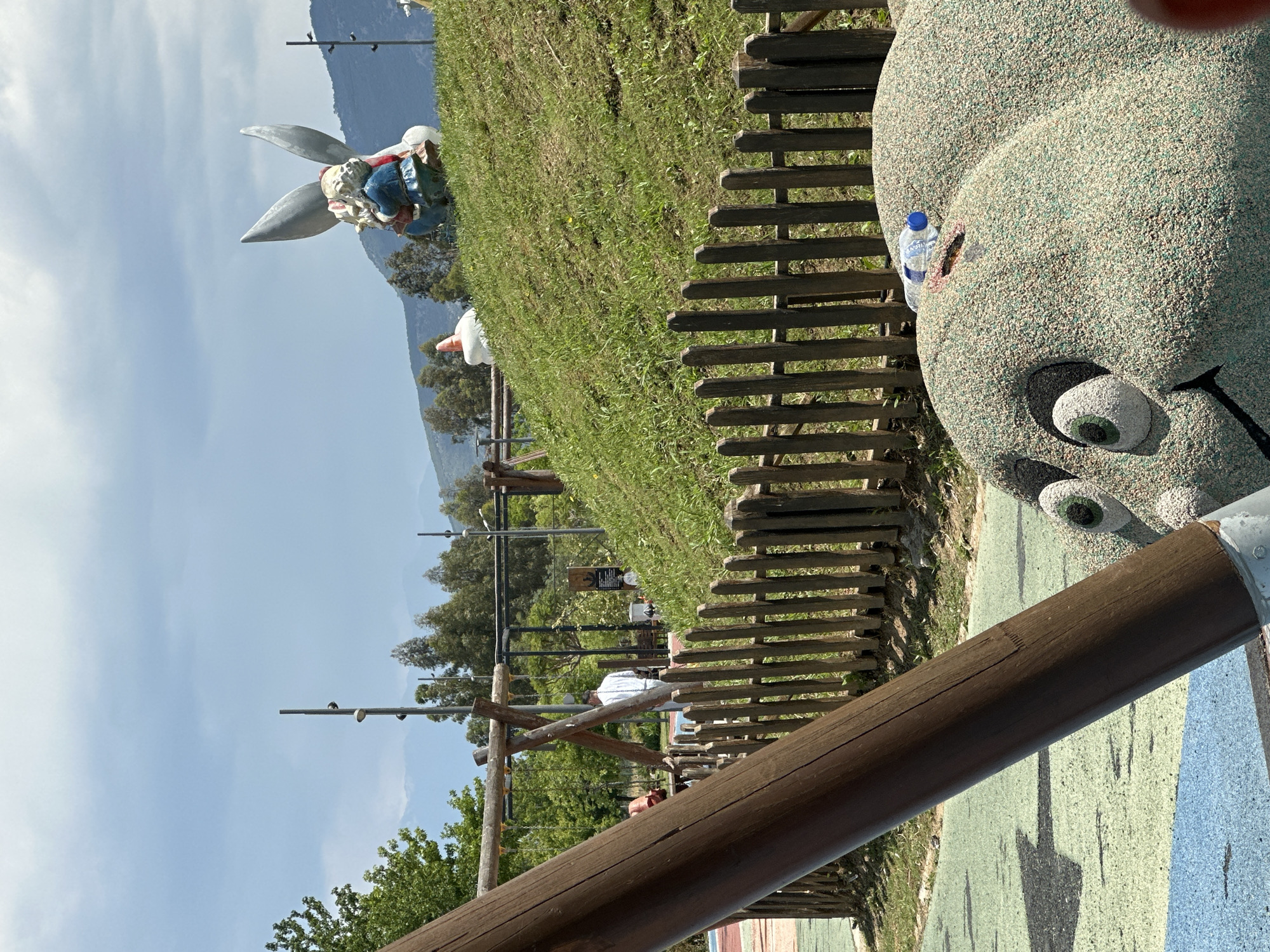 The image shows a landscape in the city of Fethiye, Turkey, featuring a playground with various attractions and a scenic backdrop of mountains under a sky with scattered clouds.
The playground features a large, textured statue of a head or animal figure with prominent eyes, positioned next to a wooden fence. A water bottle is placed near the head. In the background, there is a statue of a figure with rabbit ears, dressed in clothing.
The playground also includes wooden beams and structures, forming what appears to be climbing equipment. Trees and green foliage dot the scene, adding to the natural environment. There are people present in the background. The ground has a painted surface with various colors, predominantly green and blue.