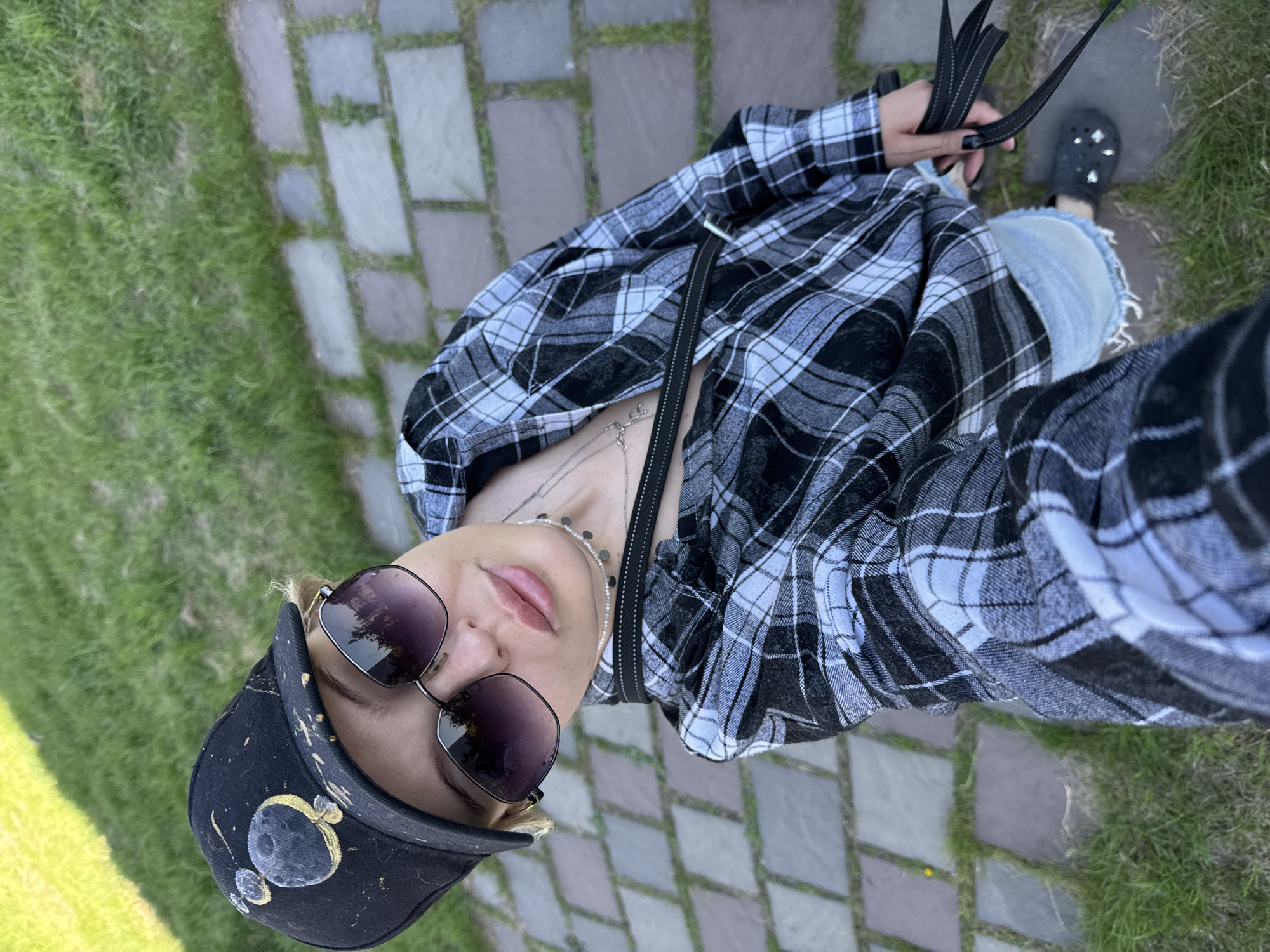 A high-angle, diagonally oriented selfie captures an individual outdoors in Krasnoyarsk, Russia. The person, viewed from the chest up, is looking directly at the camera with a neutral expression, partially obscured by dark, square-framed sunglasses that reflect what appears to be trees or an overcast sky. They wear a black and white plaid shirt, unbuttoned to reveal multiple silver chain necklaces, including one with small, flat, disc-like charms.
On their head is a black visor or baseball cap, featuring a unique, possibly hand-painted design of two moon-like celestial bodies with gold splatters. The person's right hand is visible, holding a black bag strap, and they have dark nail polish. Below the shirt, light-wash ripped jeans are visible, with a tear showing skin on the knee. A black clog-style shoe, resembling a Croc, adorned with shiny decorative elements, is visible on the foot in the background.
The setting is an outdoor area with a ground composed of interspersed patches of vibrant green grass and irregular, light-colored paving stones. The lighting suggests it is daytime, possibly late afternoon or early evening. The overall scene conveys a casual and relaxed atmosphere. No visible text is present in the image.