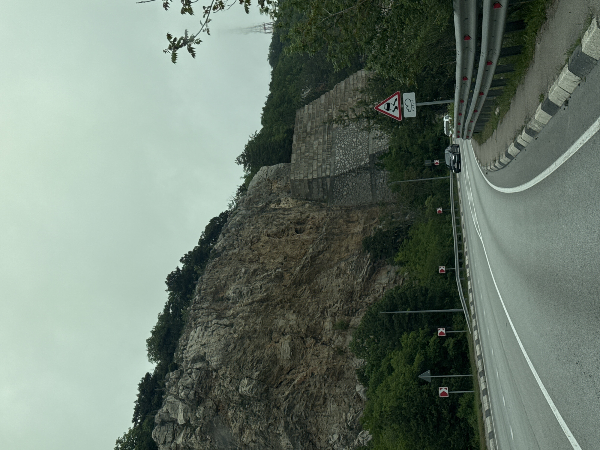 A road winds along a cliff face in Симеизский поселковый совет, Ukraine. A grey sky hangs above. A stone wall supports the cliff face. A guardrail runs along the road's edge, punctuated by reflective markers. A road sign warns of slippery conditions and crosswinds. Two cars are parked on the roadside. Turn markers are visible. The cliff face is covered with vegetation.