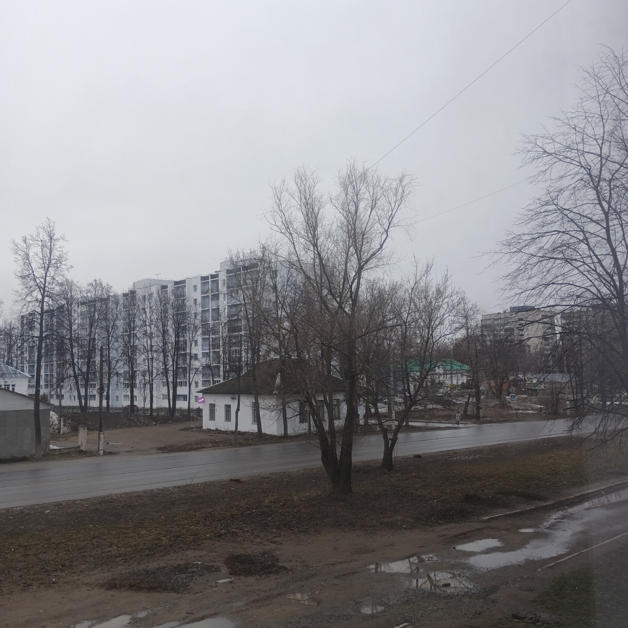 The image depicts a street scene in Chaikovsky, Russia on a gray, overcast day. The street is wet and reflective, suggesting recent rain. A brown, muddy area with puddles is in the foreground. A two-lane road runs horizontally through the middle of the scene. On the other side of the road is a strip of land with trees lacking leaves and a few small buildings. A white, single-story building with a dark green roof and a pink sign is visible. Behind the small buildings, a large, modern apartment building is seen. The sky is uniformly gray, and a thin wire is stretched across the upper portion of the image.