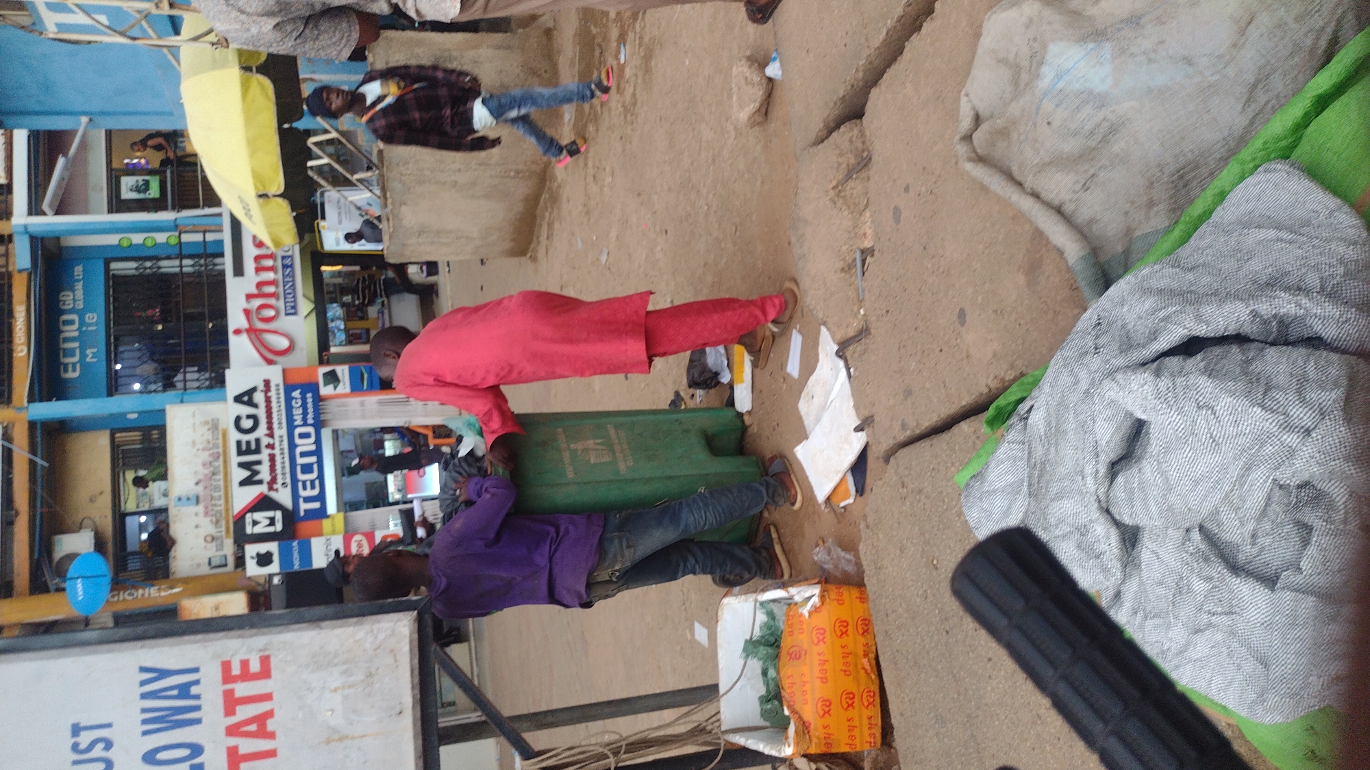 A street scene in Jos, Nigeria, depicts two men working on or near a green trash receptacle. One man, wearing a red outfit, bends over the container, while the other, in a purple shirt, is on the opposite side. Several business signs are visible in the background, including "ECNO GD GLOBAL LTD," "John's PHONES & C," "MEGA Phones & Accessories," "TECNO MEGA," and "GIONEE." Other subjects in the background are a person walking in a plaid shirt and a person in a gray top. A pile of material sits on the ground, along with a discarded orange box labeled "RX shep." The environment is a bustling urban setting.
